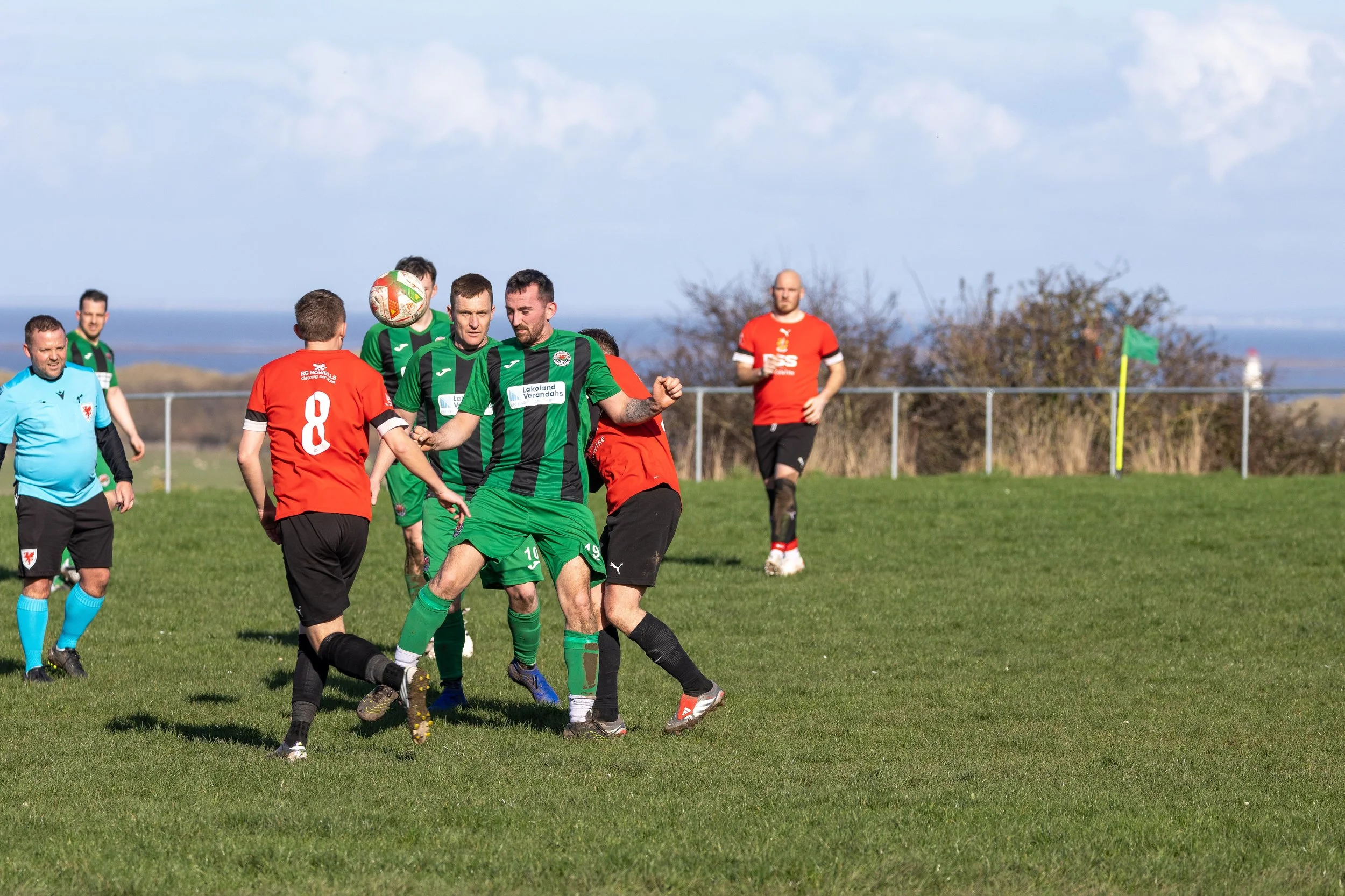 Soccer players competing for the ball on a green field with a fence and trees in the background under a partly cloudy sky.