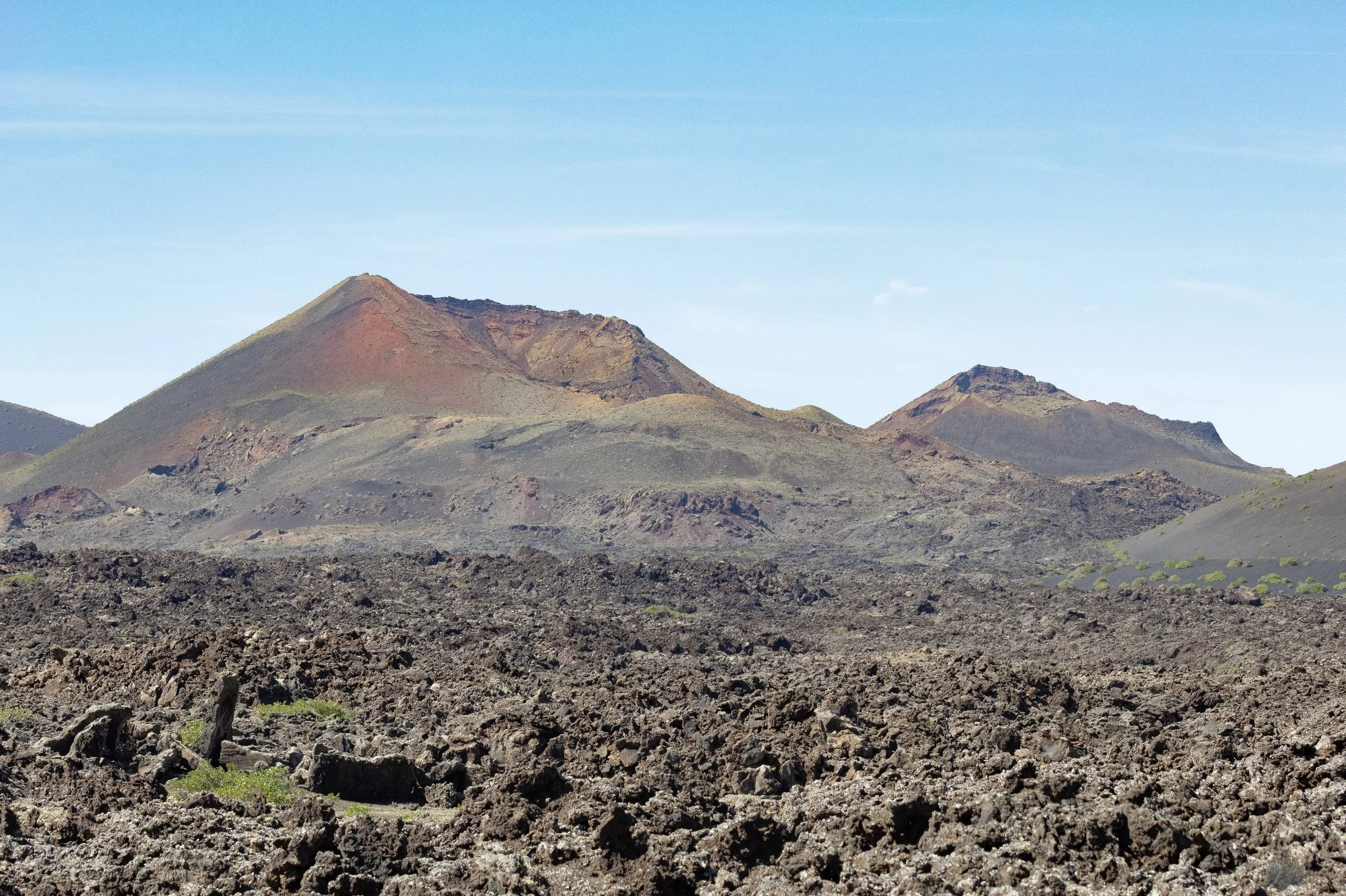 A volcanic landscape with rugged, dark lava rocks in the foreground and distant volcanoes with sloped sides against a clear blue sky.