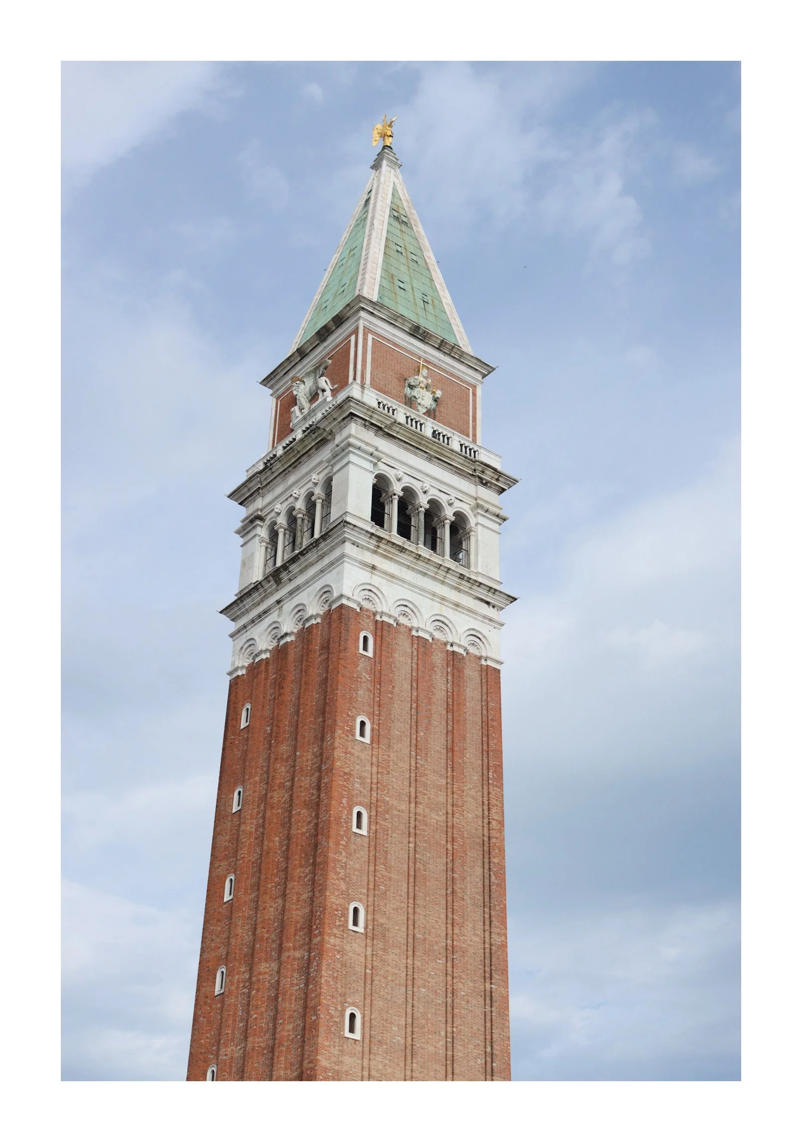 A tall brick bell tower with a green roof and golden statue on top, set against a partly cloudy sky.