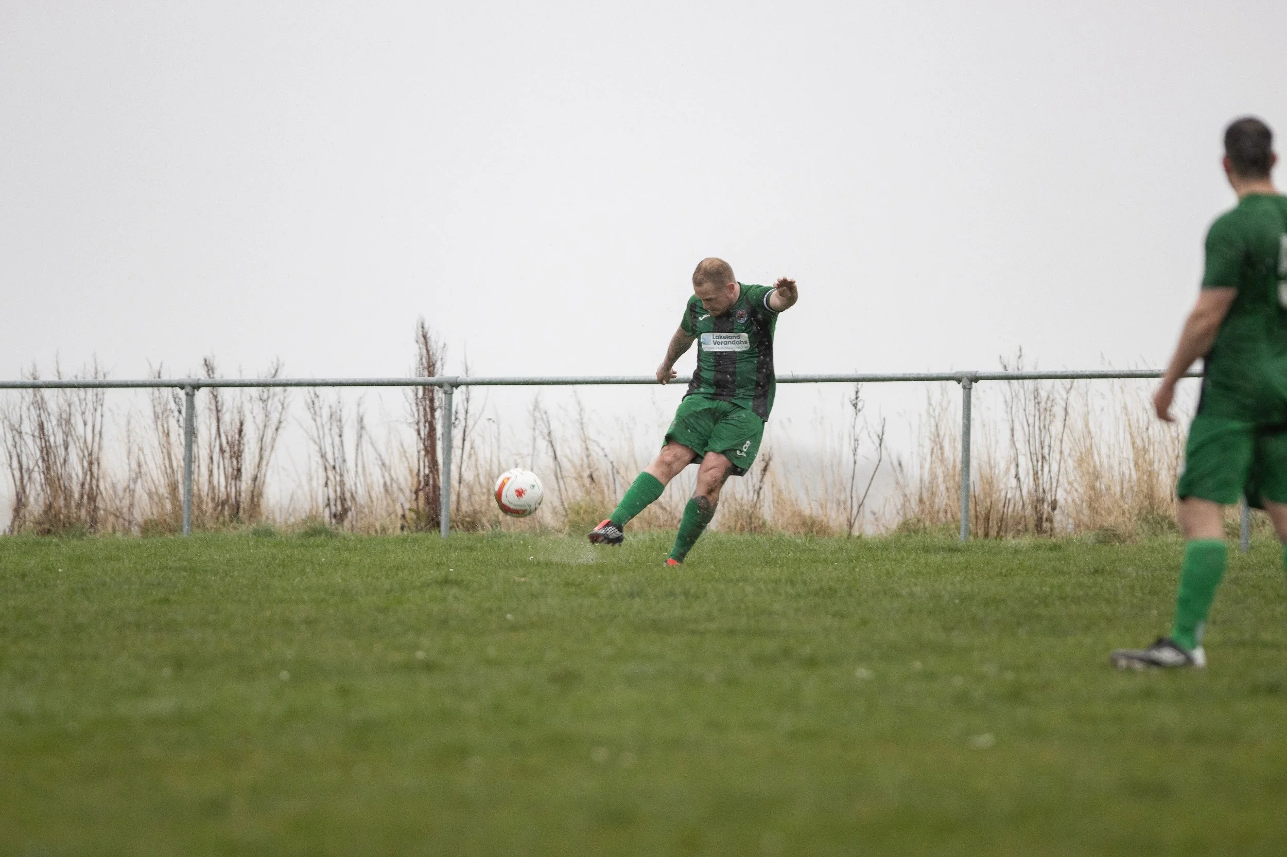 A soccer player in a green and black uniform kicking a white and orange soccer ball on a grassy field. Another player in a similar uniform is partially visible on the right. The background shows a metal fence and dry grass under an overcast sky.