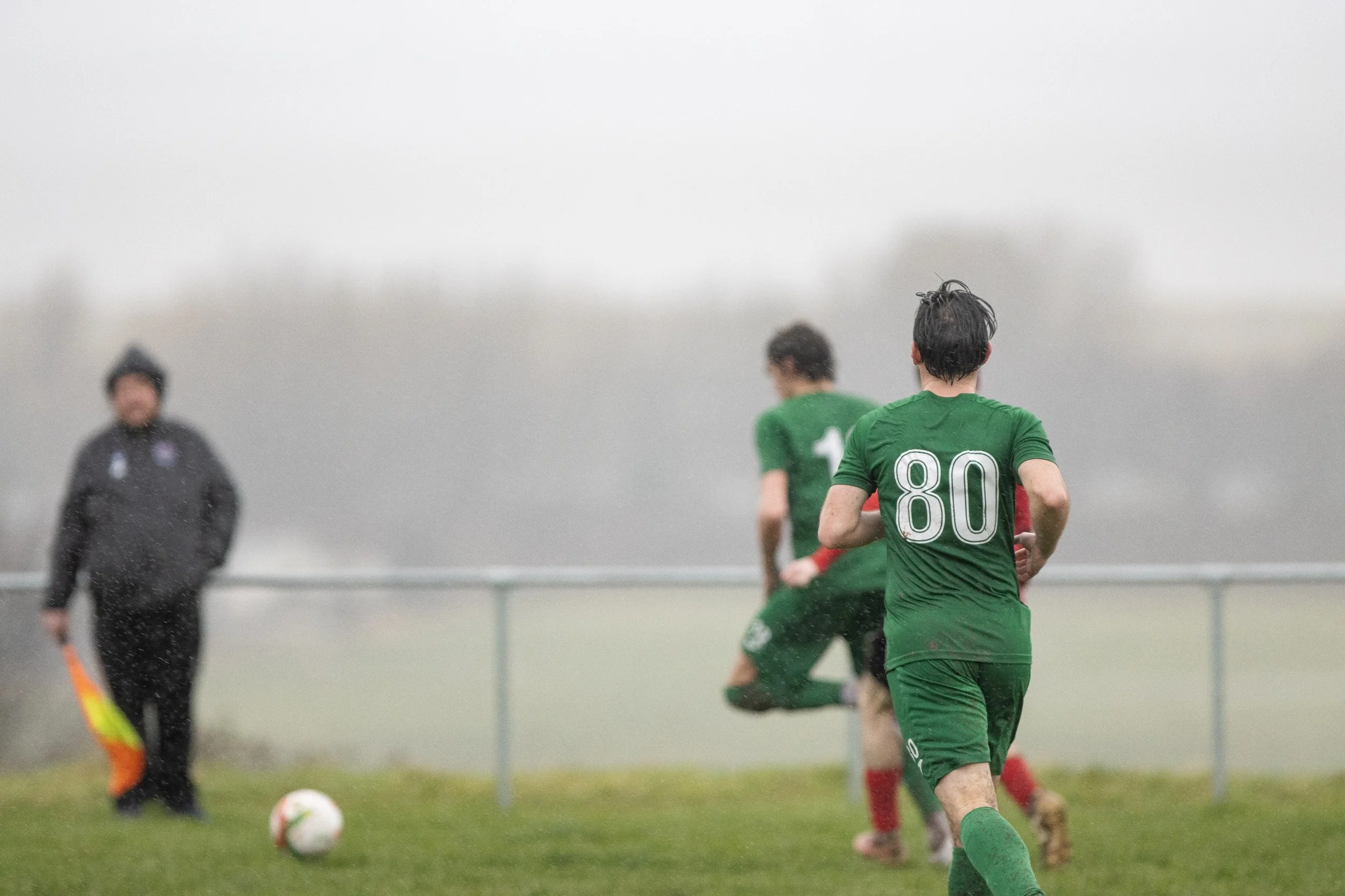 Soccer players in green jerseys on a rainy field with an official in black holding a flag, in an outdoor game under cloudy sky.