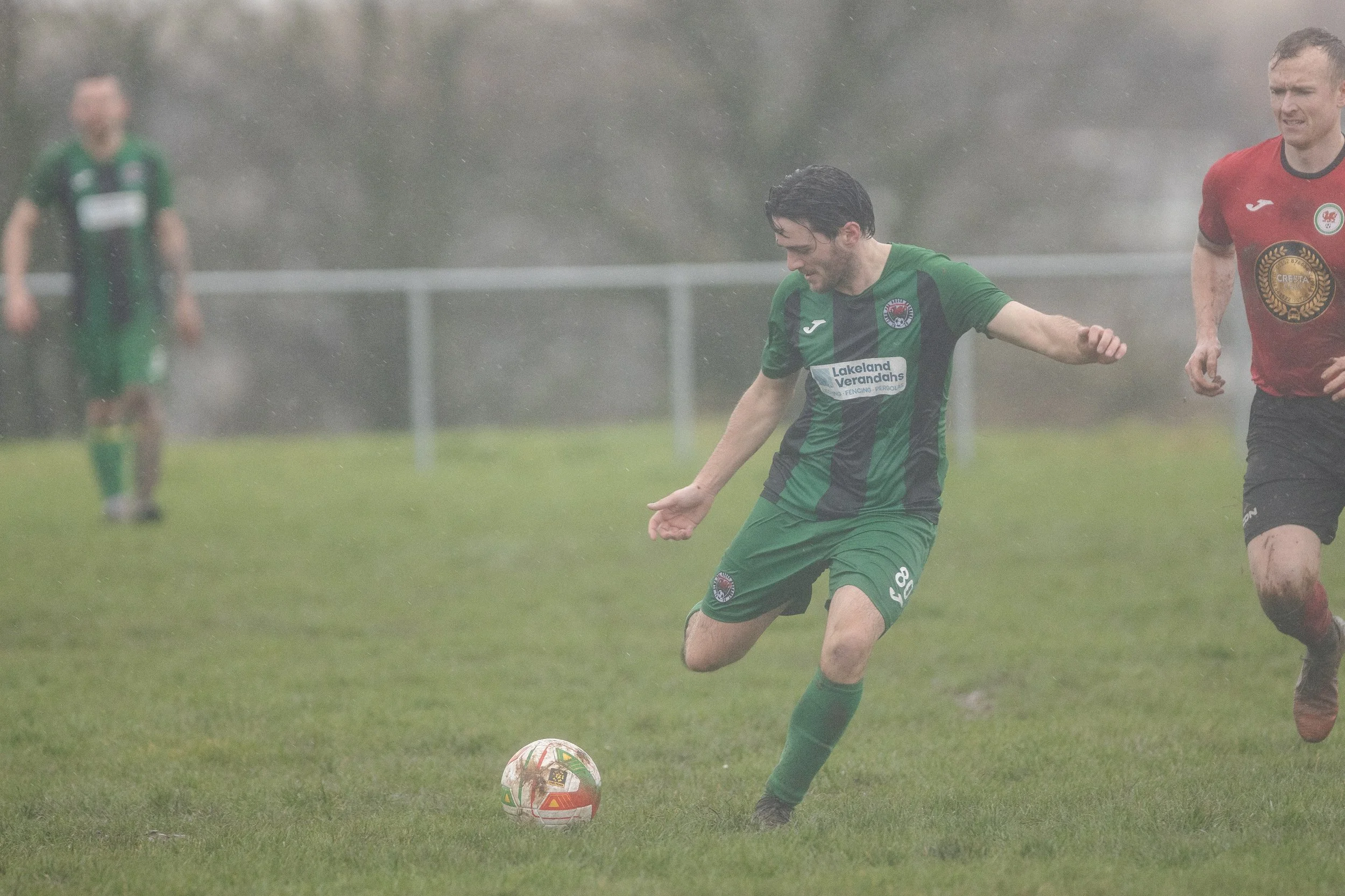 Soccer players in rain, green and red jerseys, one kicking ball on wet grass field.