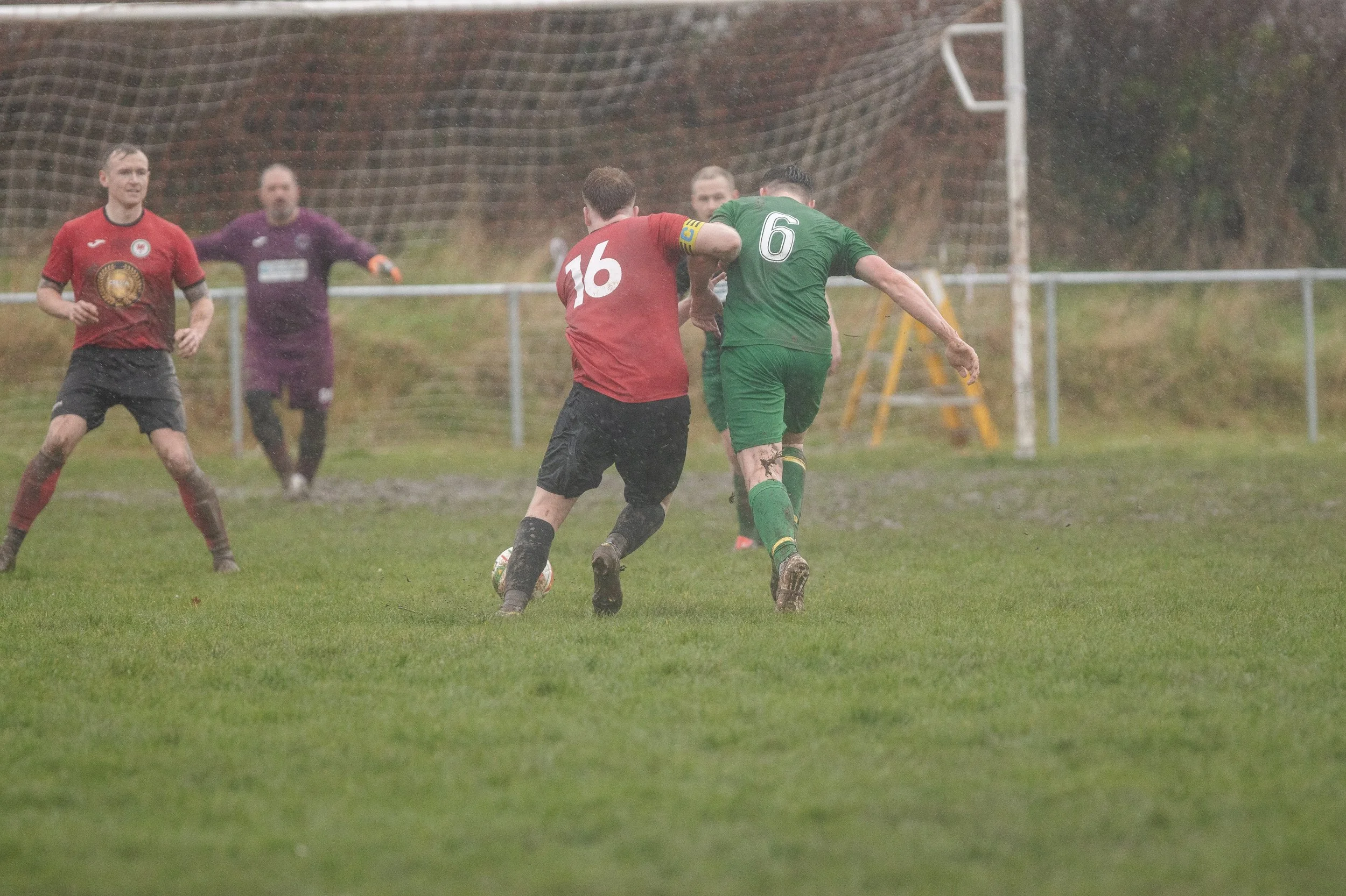 Soccer players in red and green jerseys competing for the ball on a muddy field in the rain.