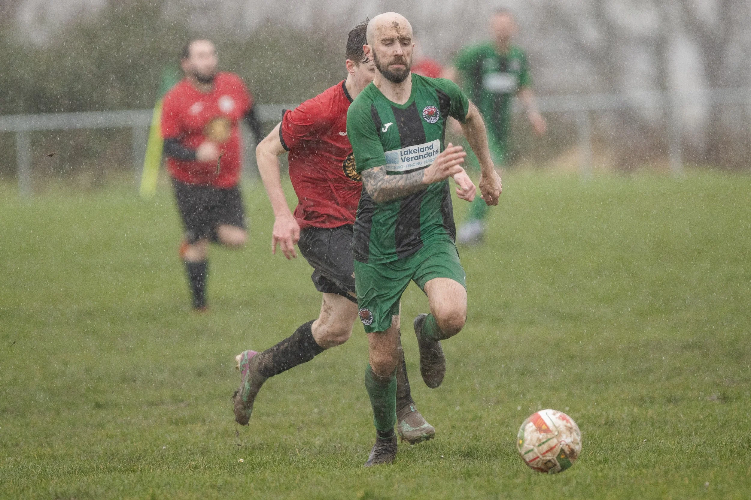 A soccer game in the rain with players running after the ball on a grassy field, one player in a green and black uniform leading with the ball.