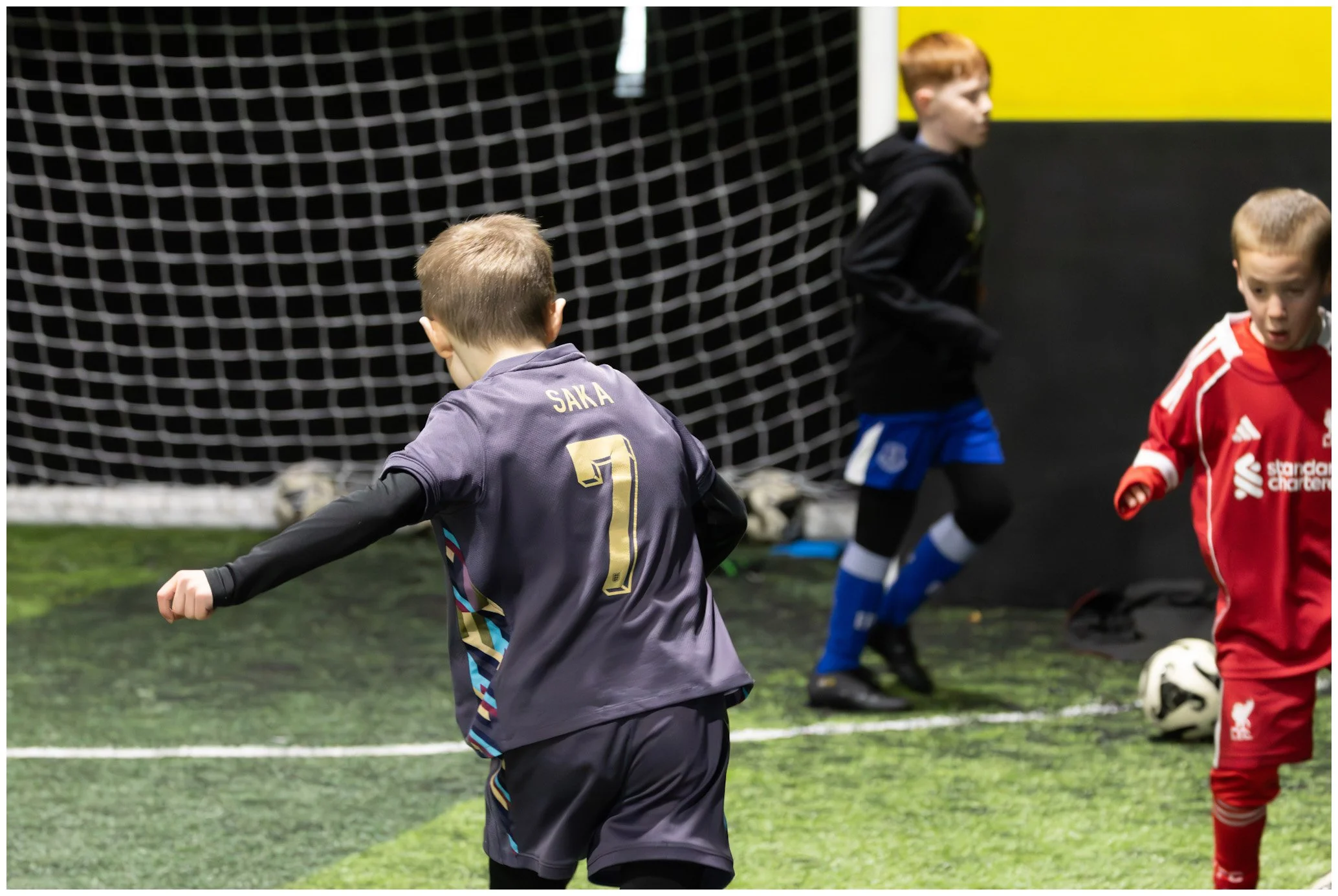 Kids playing indoor soccer, with one in a gray goalkeeper jersey with the name Saka, and others in black and red jerseys, near a goal with a net.