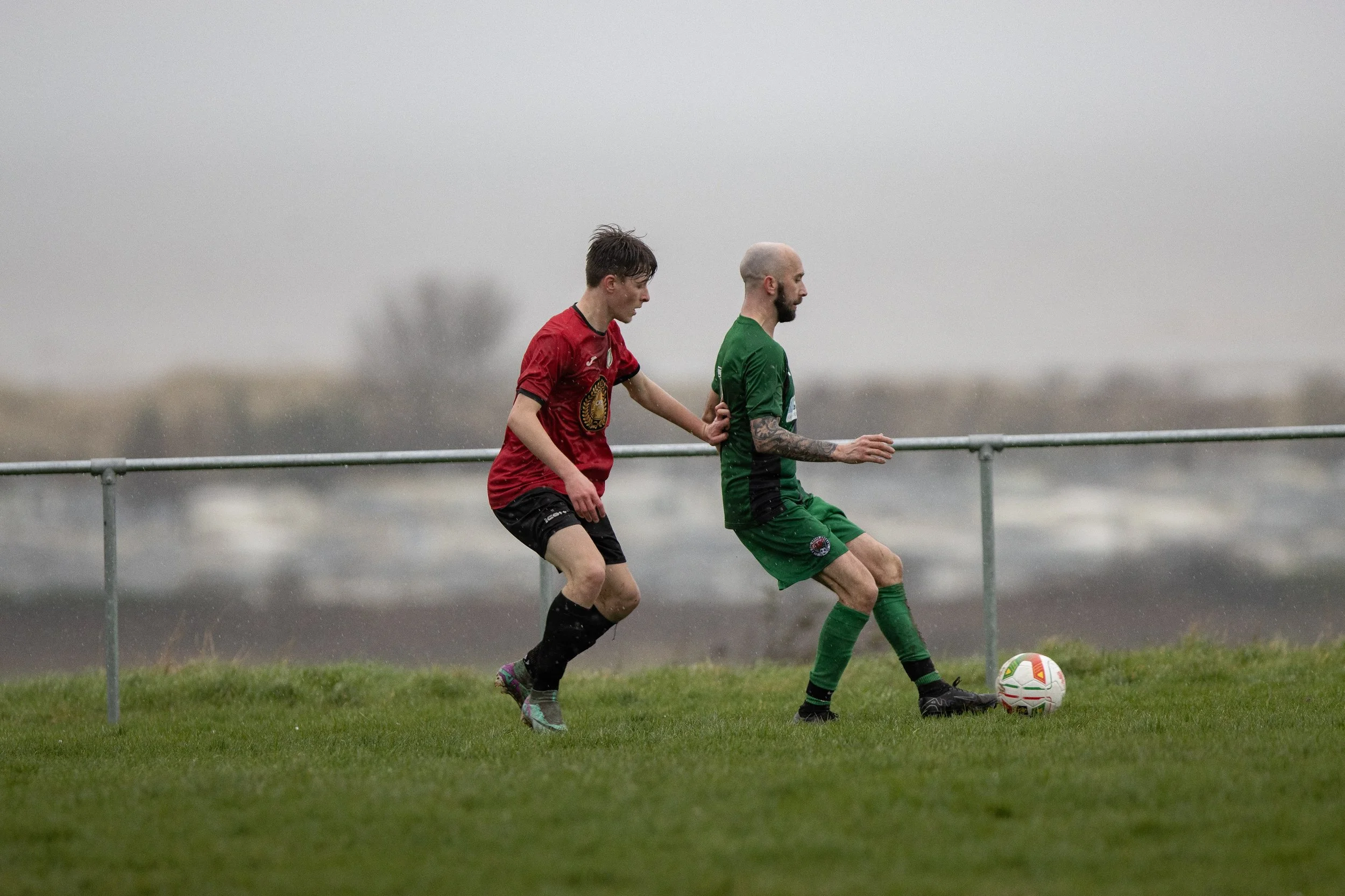 Two soccer players competing for ball on rainy day field with gray sky in background.