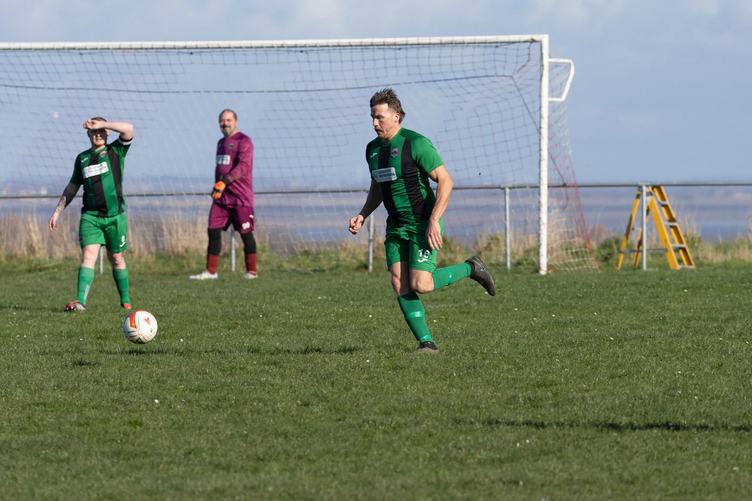 A soccer player in a green and black uniform is running on a grass field. Two other players, including a goalkeeper in a purple kit, are in the background near the goal. One of the players is standing with a ball at his feet, on a mostly cloudy day.