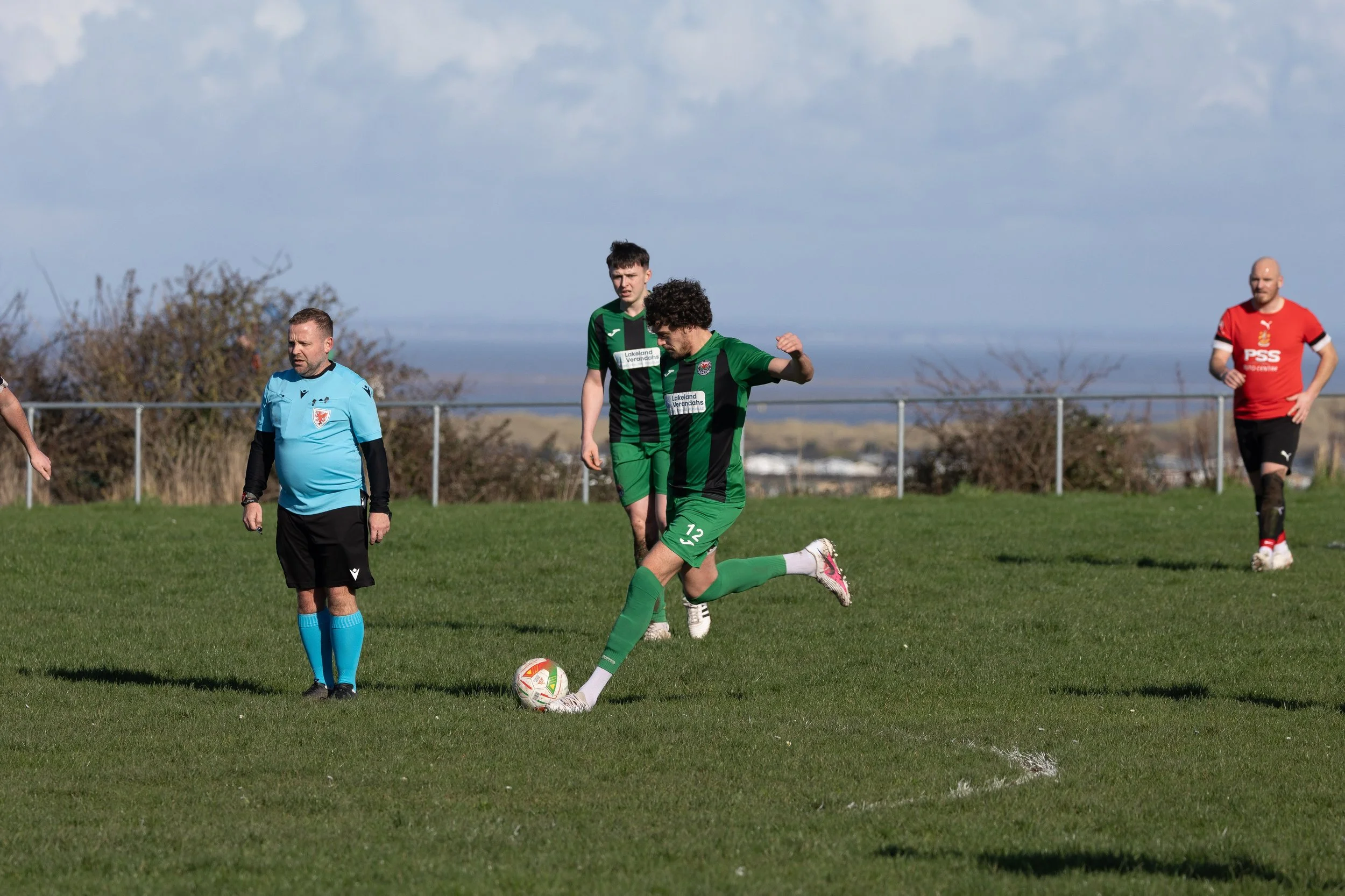 A soccer game with players on a grassy field, one player in green and black uniform about to kick the ball, a referee in a light blue shirt and black shorts nearby, and other players in the background, with a chain-link fence and hills in the distanc