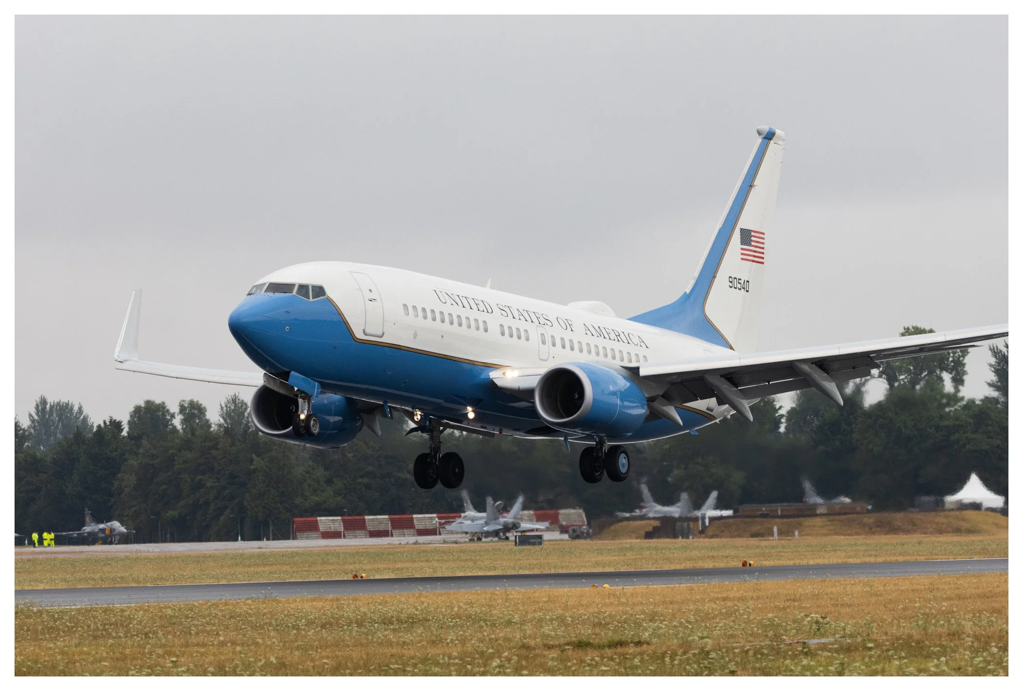 An aircraft with the U.S. Air Force livery taking off from a runway, with a cloudy sky and other planes in the background.