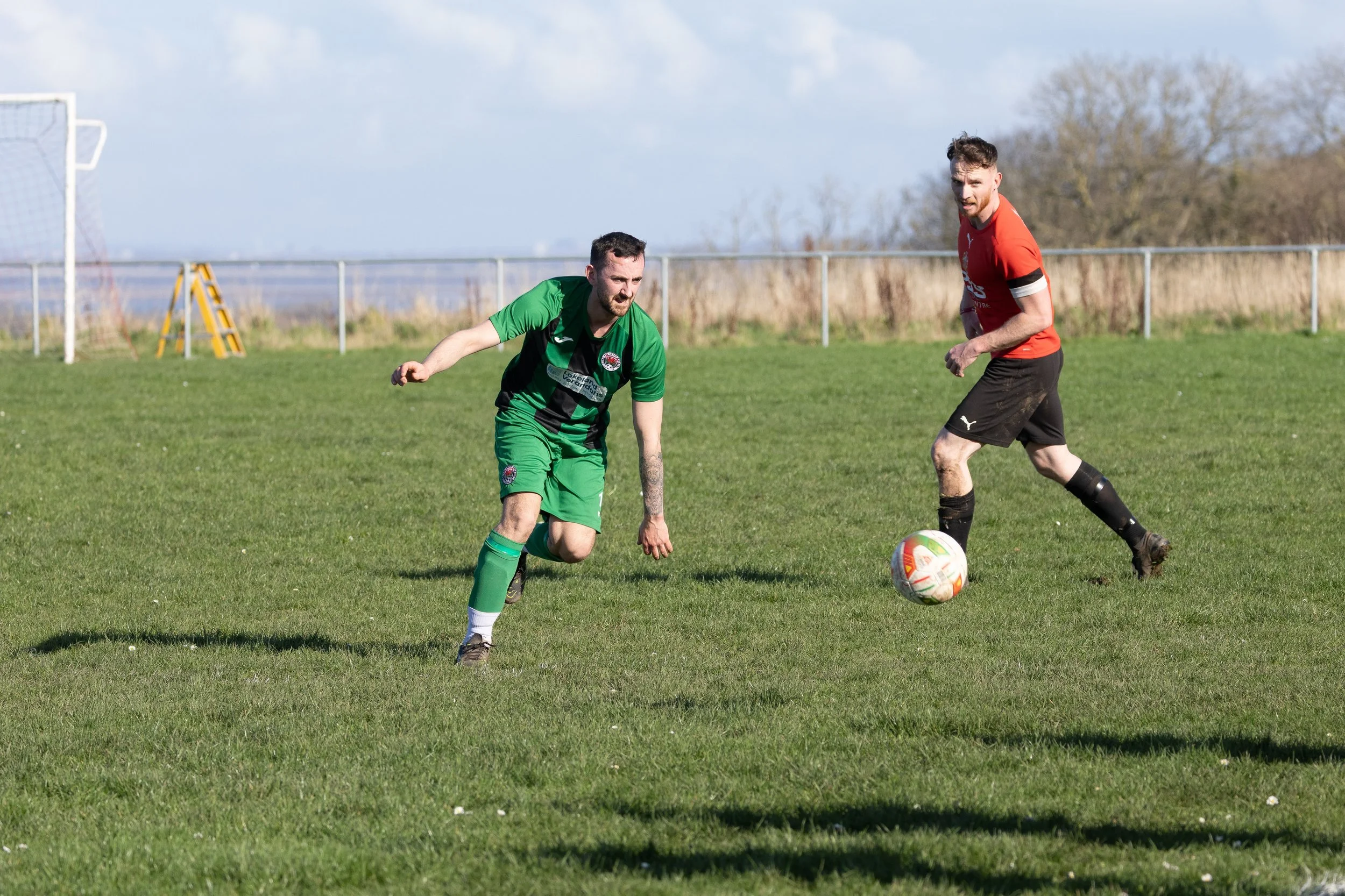 Two soccer players, one in a green and black uniform and the other in a red and black uniform, are playing on a grassy field with a soccer goal and a fence in the background.