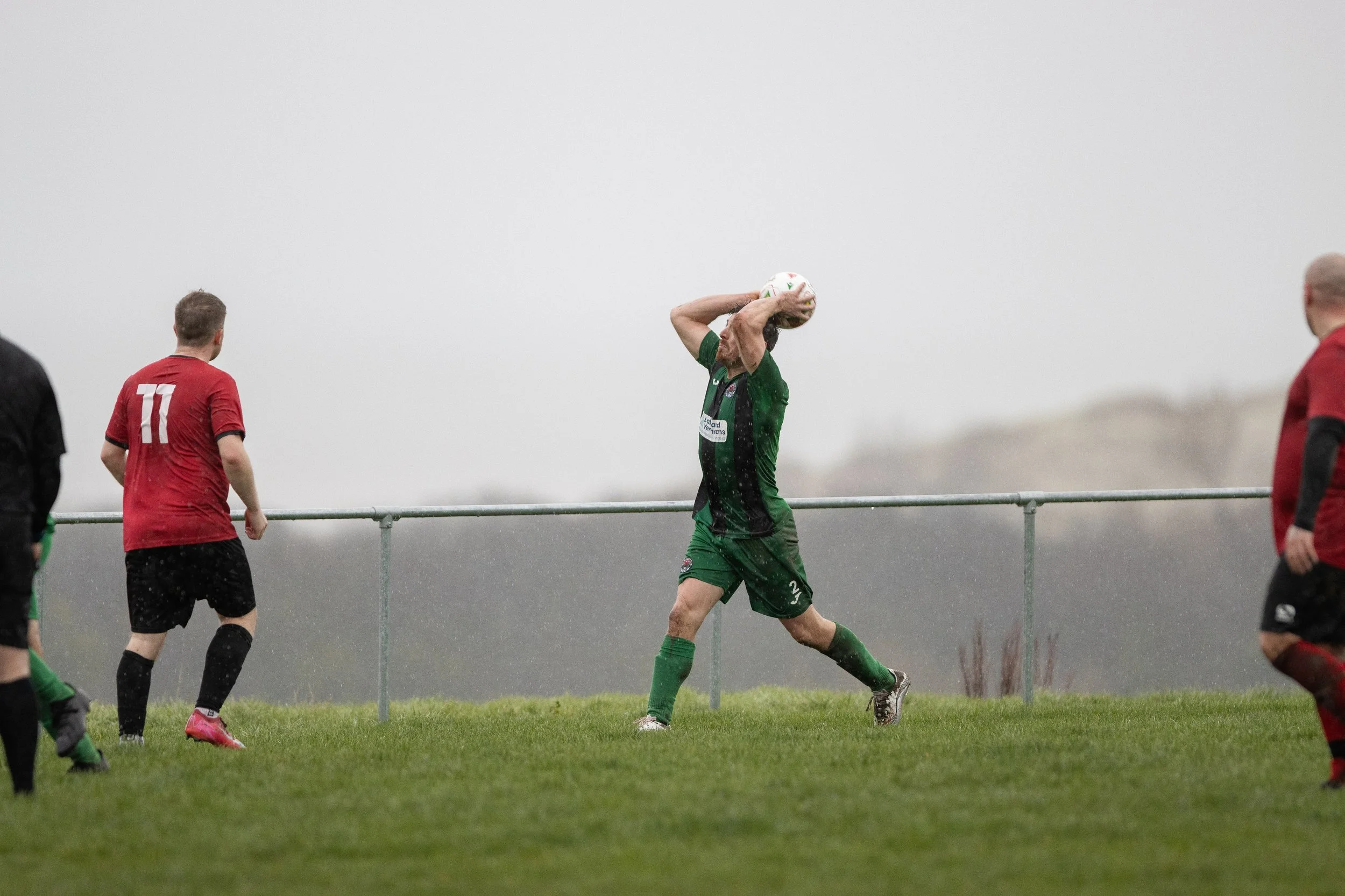 A soccer player in a green uniform is about to throw in the ball during a match on a grassy field on a rainy day with overcast skies. Other players in red and black uniforms are nearby.