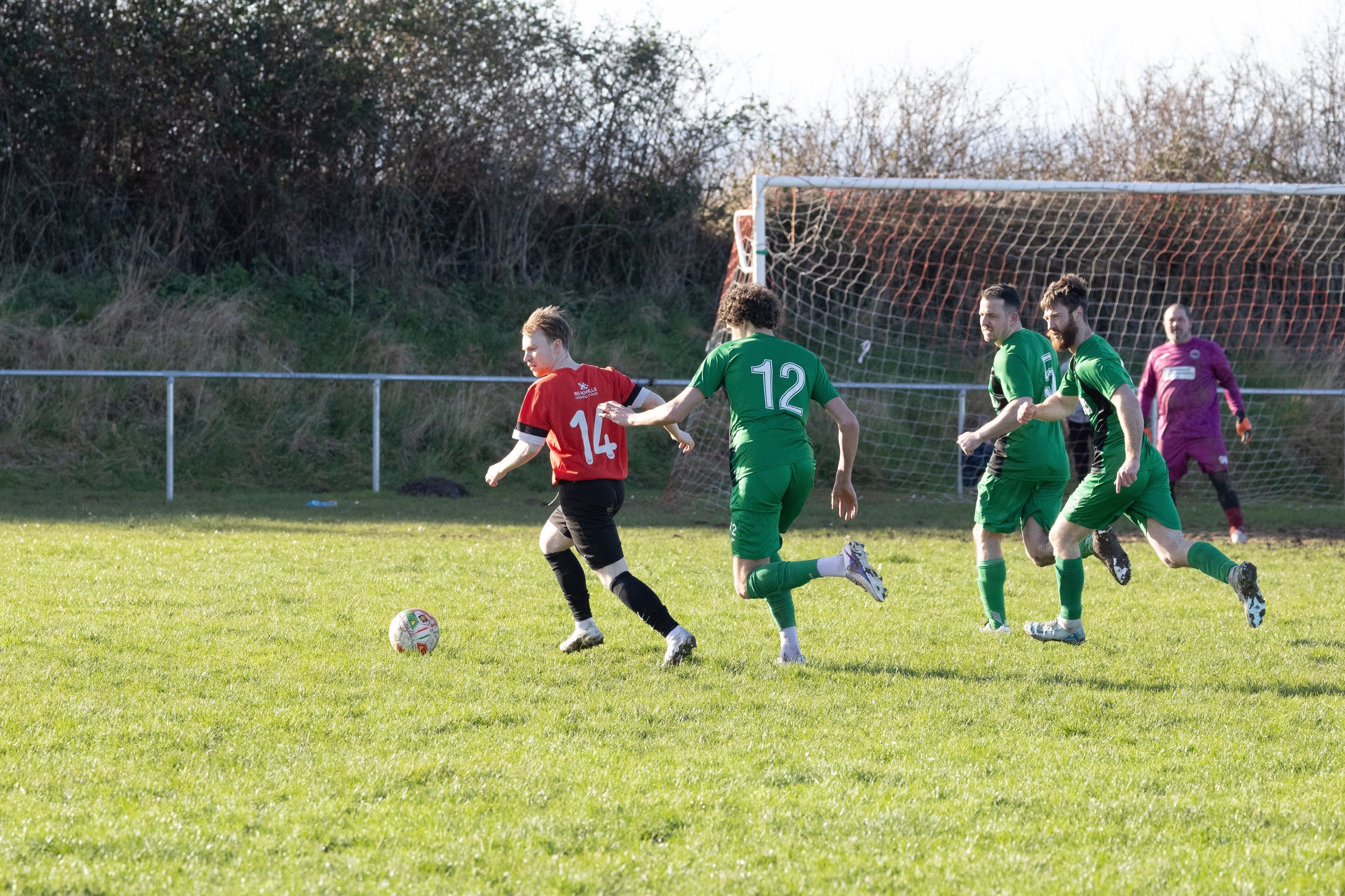 Soccer players in green and red jerseys chasing the ball on a grassy field with a goal in the background.