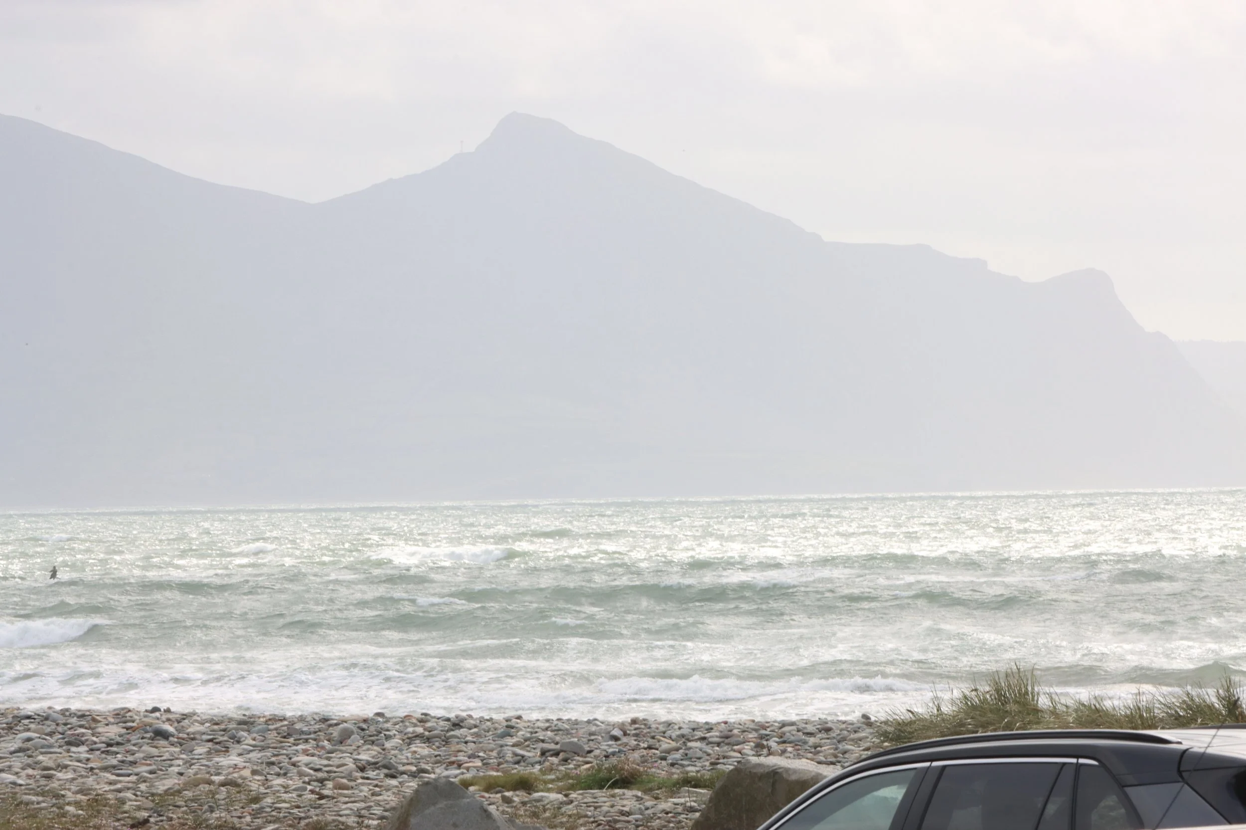 A rocky beach with waves, a car in the foreground, and a mountain in the background under a cloudy sky.