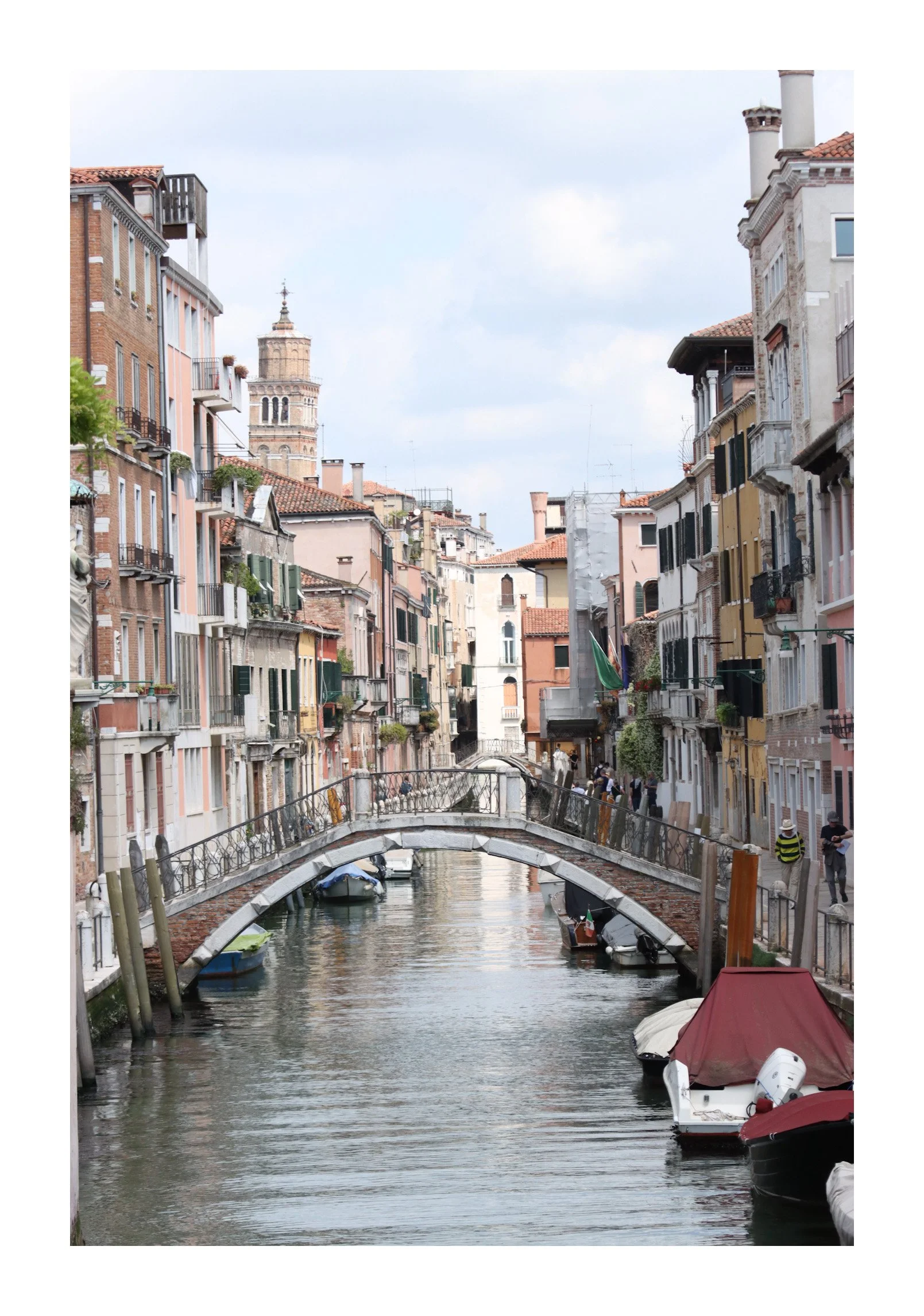 A canal scene in Venice, Italy, with boats docked along the water and colorful historic buildings lining the canal, featuring a small bridge crossing over the waterway.