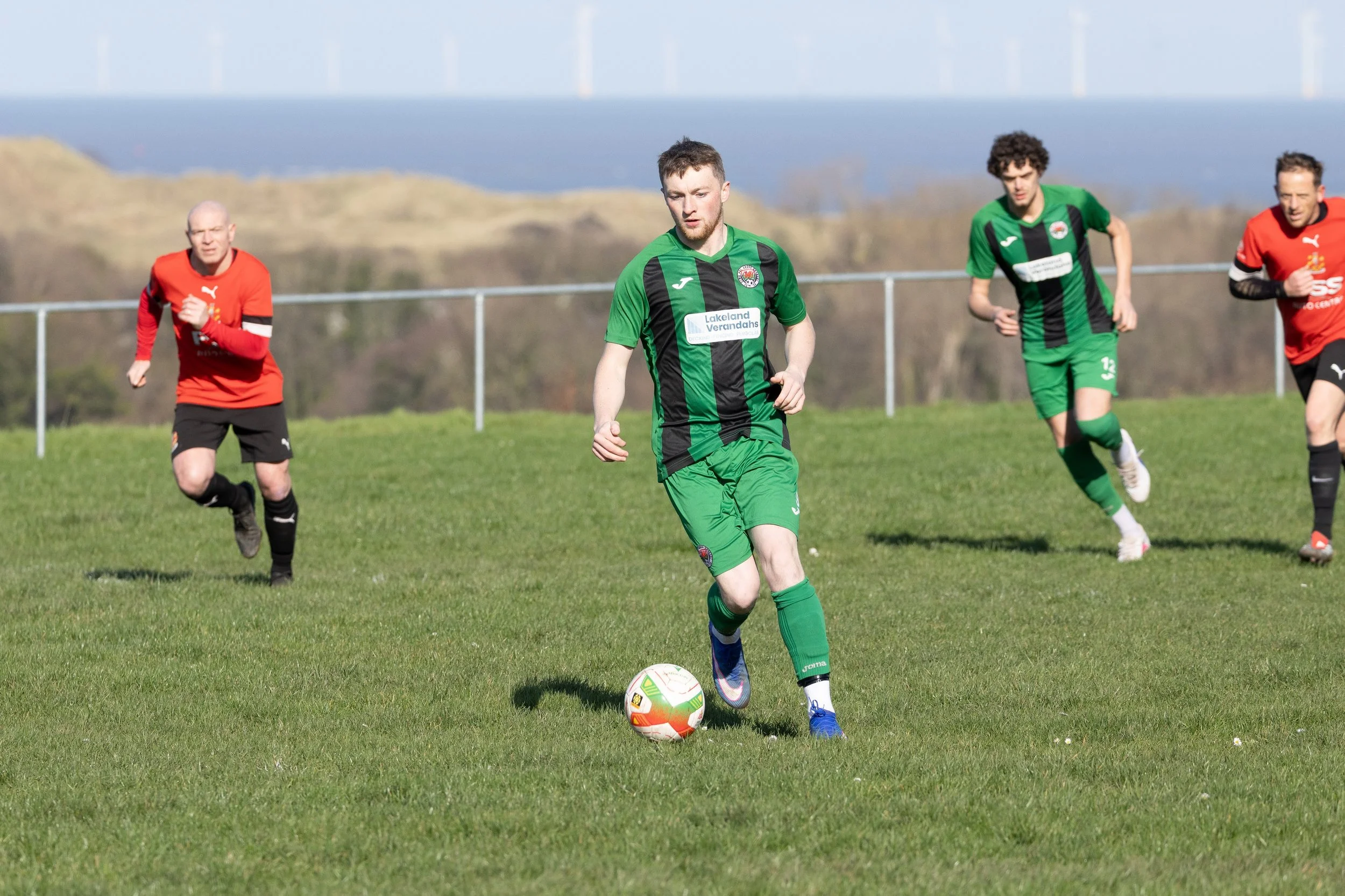 Soccer players in green and red jerseys playing on a grassy field during daytime with hills and wind turbines in the background.