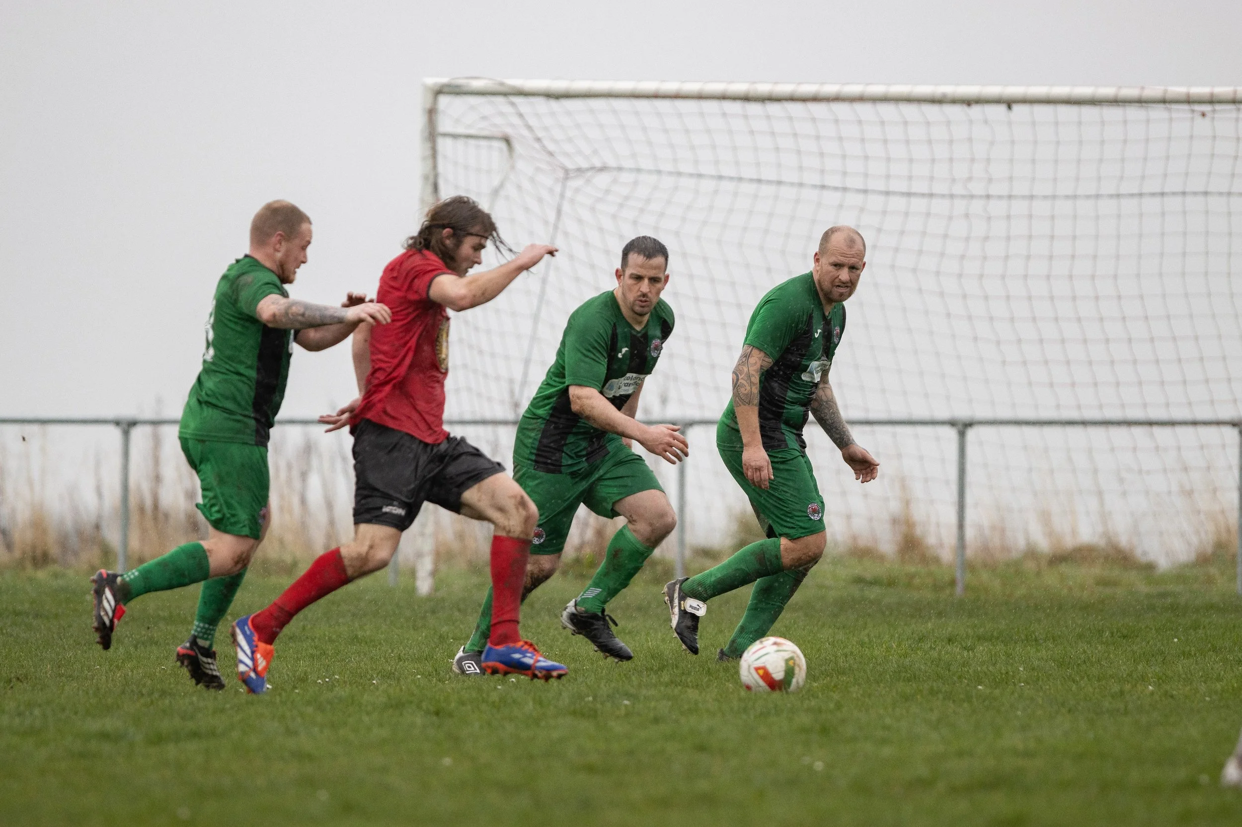 Four soccer players, three in green uniforms and one in a red uniform, are running toward the soccer ball on a grassy field near a goalpost. The scene appears to be rainy or overcast.