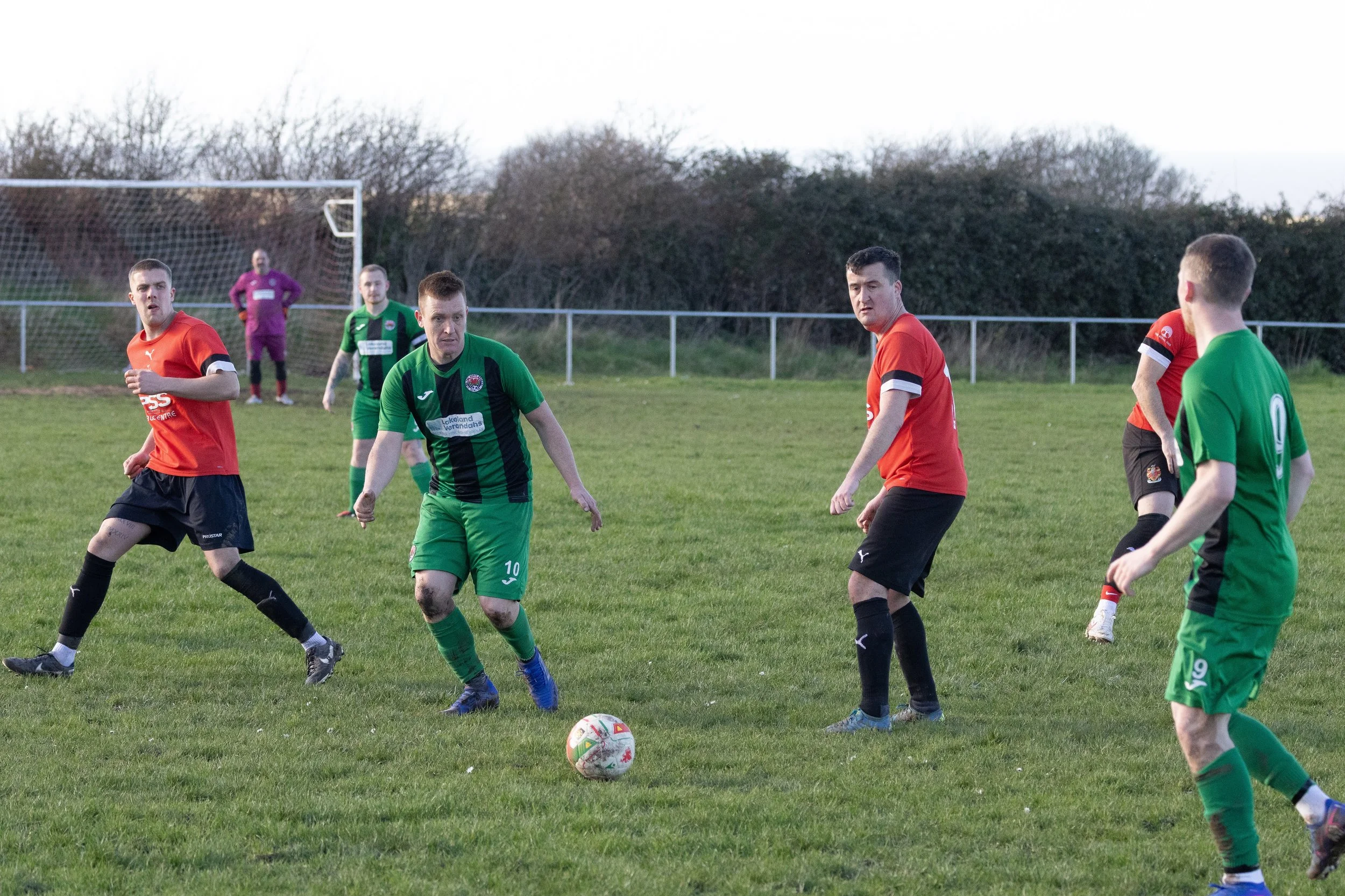 Soccer match with players in green and red jerseys on a grassy field, with a goal and trees in the background.