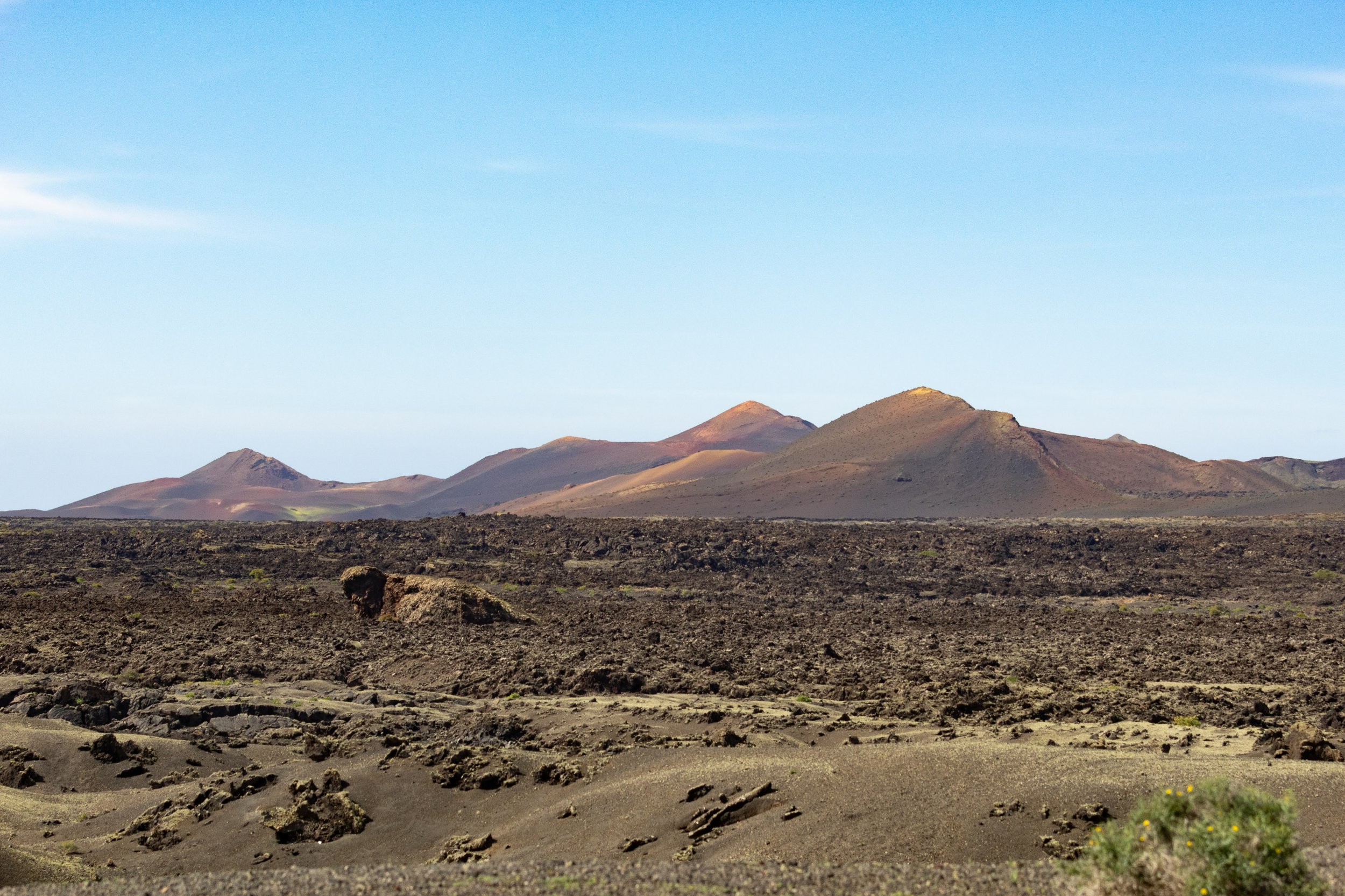 A vast desert landscape featuring dark, rocky terrain with volcanic hills and mountains in the background under a clear blue sky.
