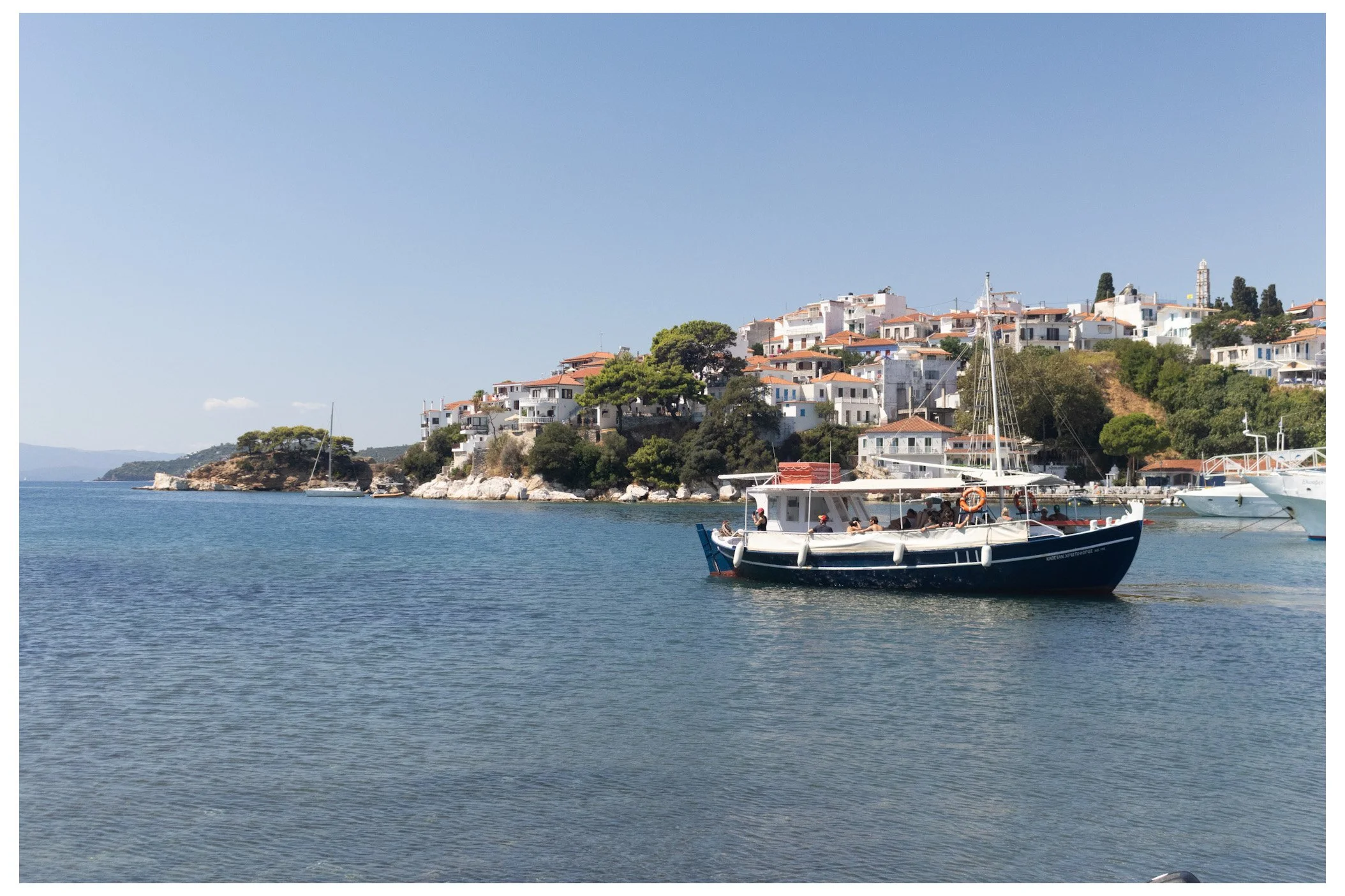 A boat floating on calm water near a coastal town with white buildings and red-tiled roofs, green trees, and a clear blue sky.