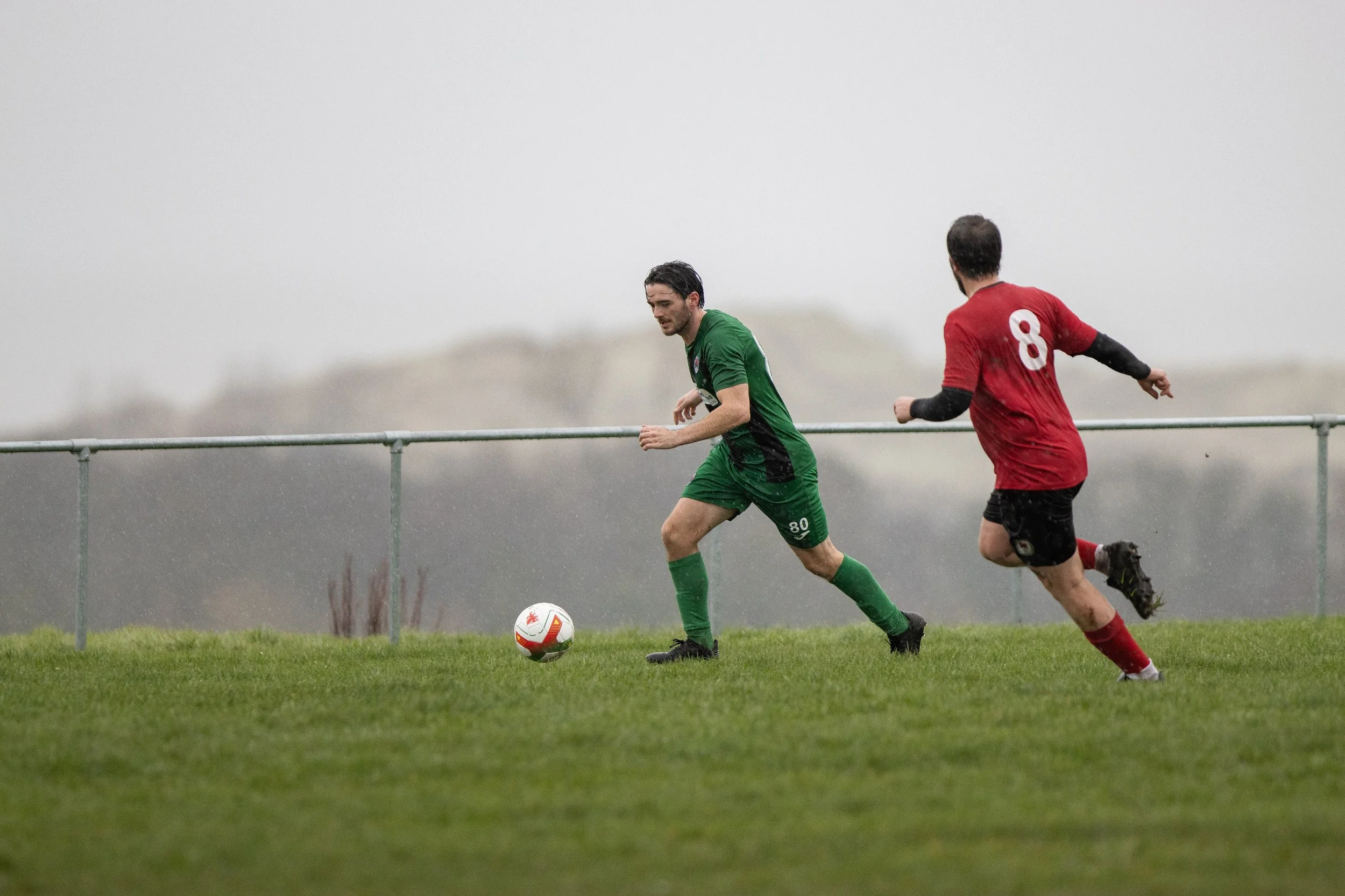 Two soccer players, one in a green uniform and the other in a red uniform, chasing a soccer ball on a grassy field under a cloudy sky.