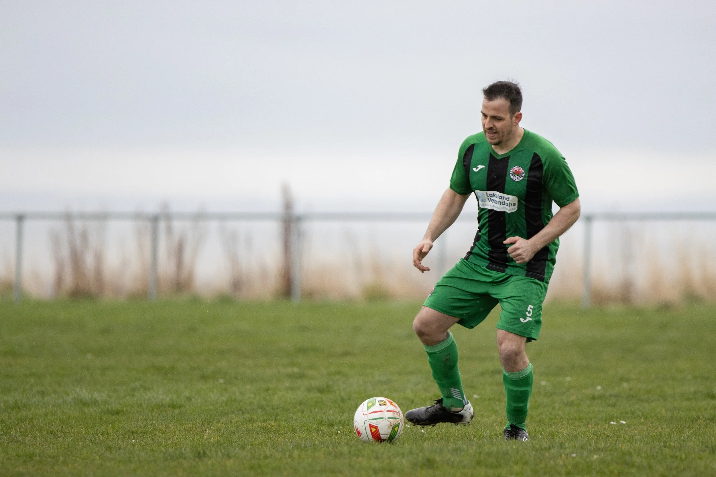 A man playing soccer on a grassy field, wearing a green and black striped jersey, green shorts, and black cleats, with a soccer ball at his feet, under an overcast sky.