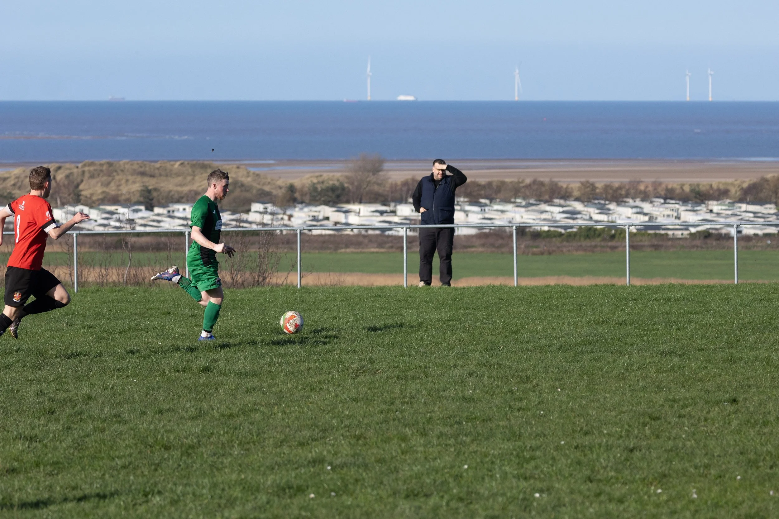 Two young men playing soccer on a grassy field with another person watching, across a coastline with wind turbines in the background.