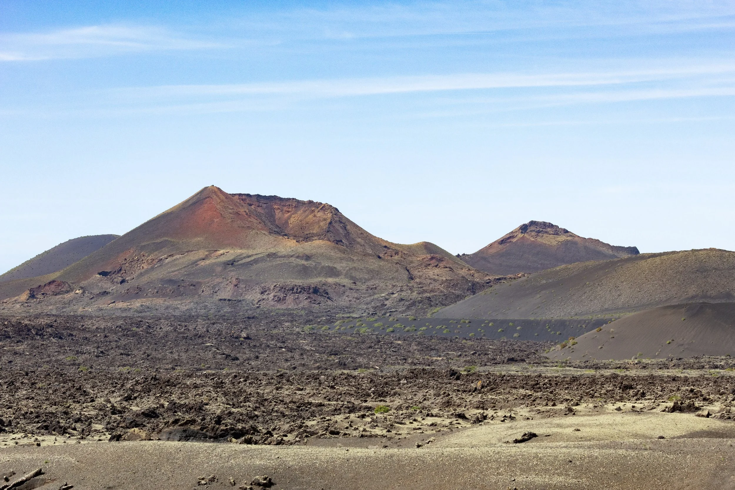 A volcanic landscape with dark, rocky terrain, and several mountains or volcanoes in the background under a blue sky with wispy clouds.