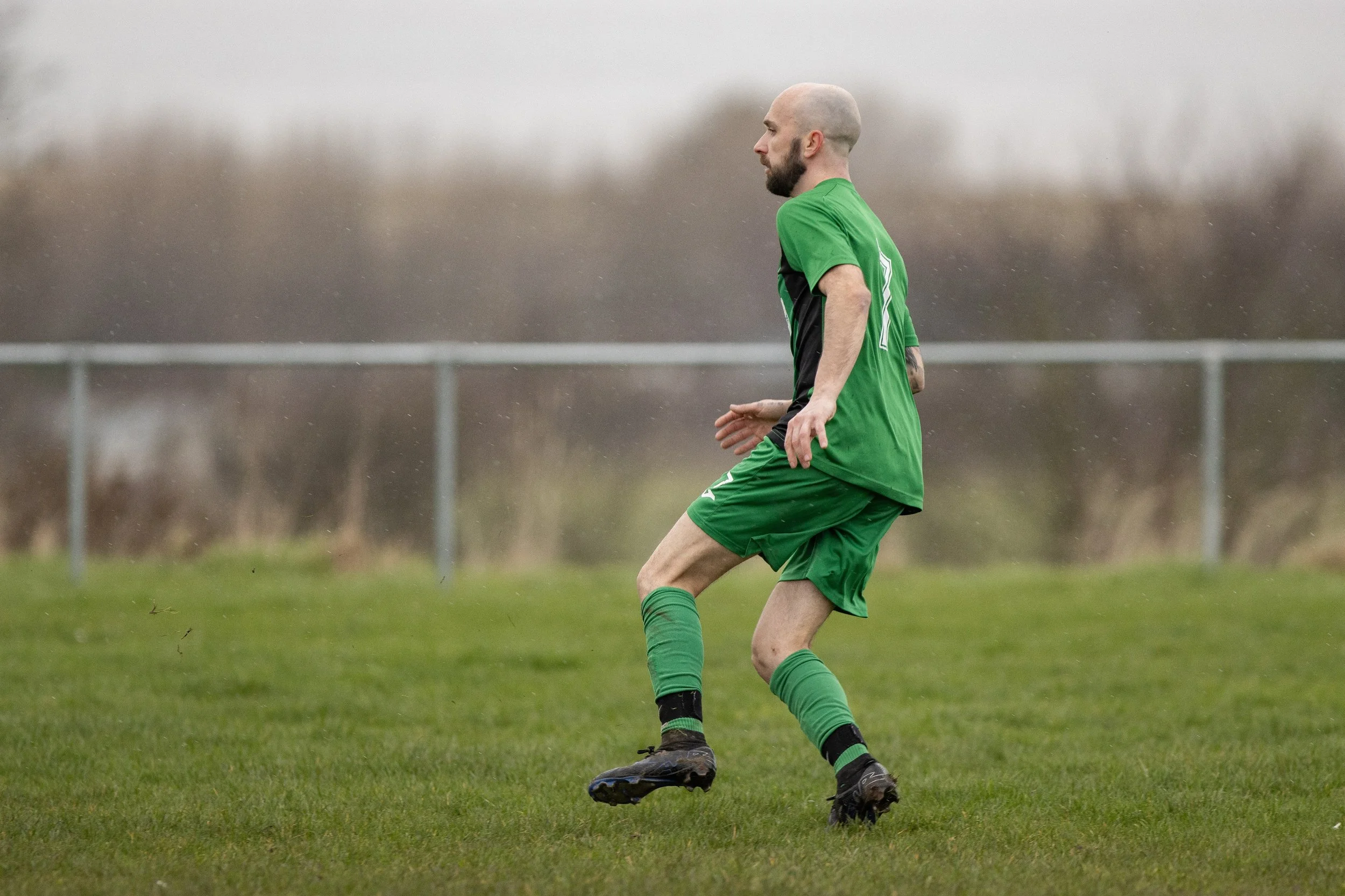 A soccer player in a green jersey and shorts kicks the ball on a grassy field, with what appears to be rain or mist falling in the background.