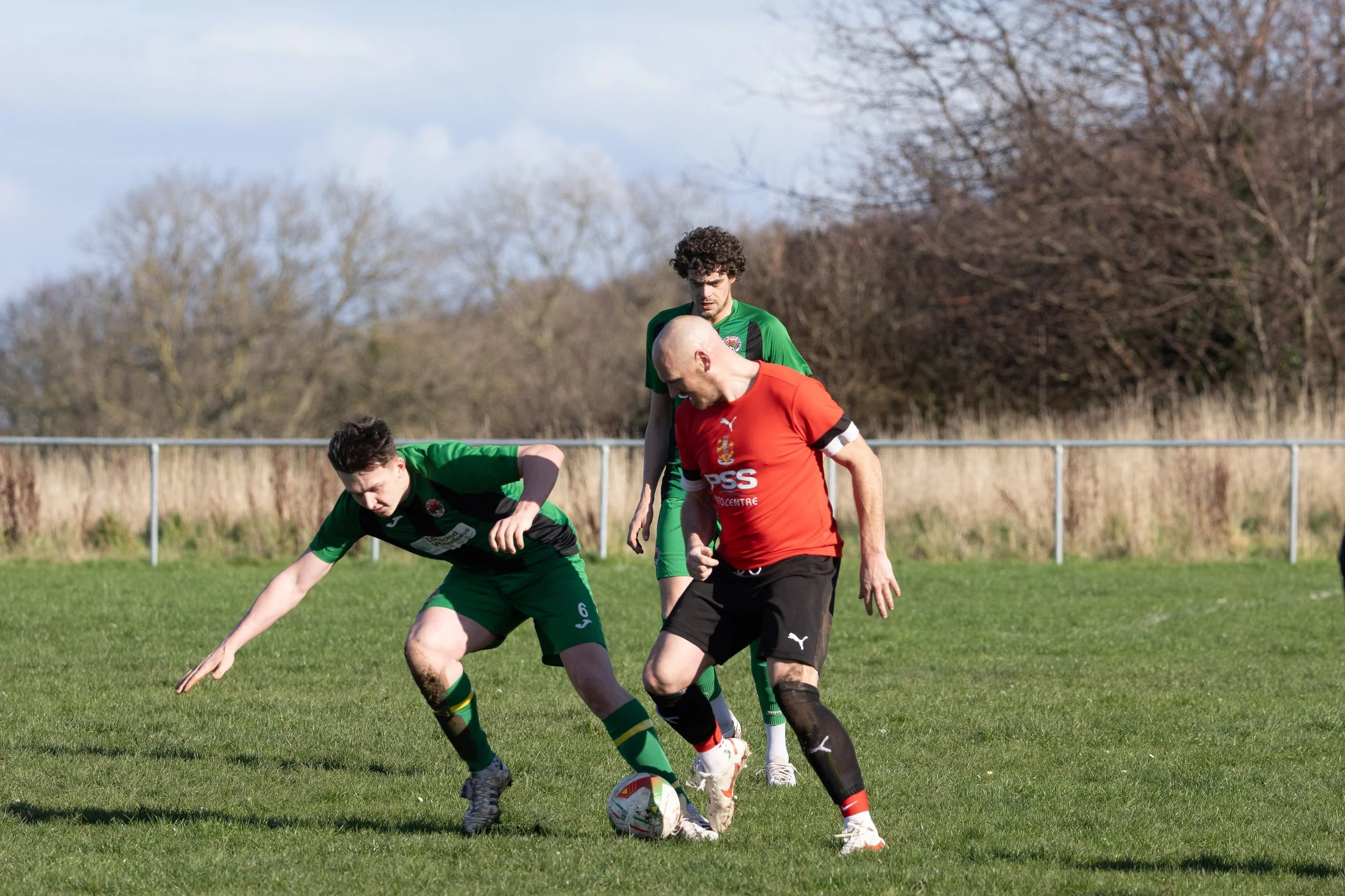 Three soccer players compete for the ball on a grassy field, with two players in green and one in red.