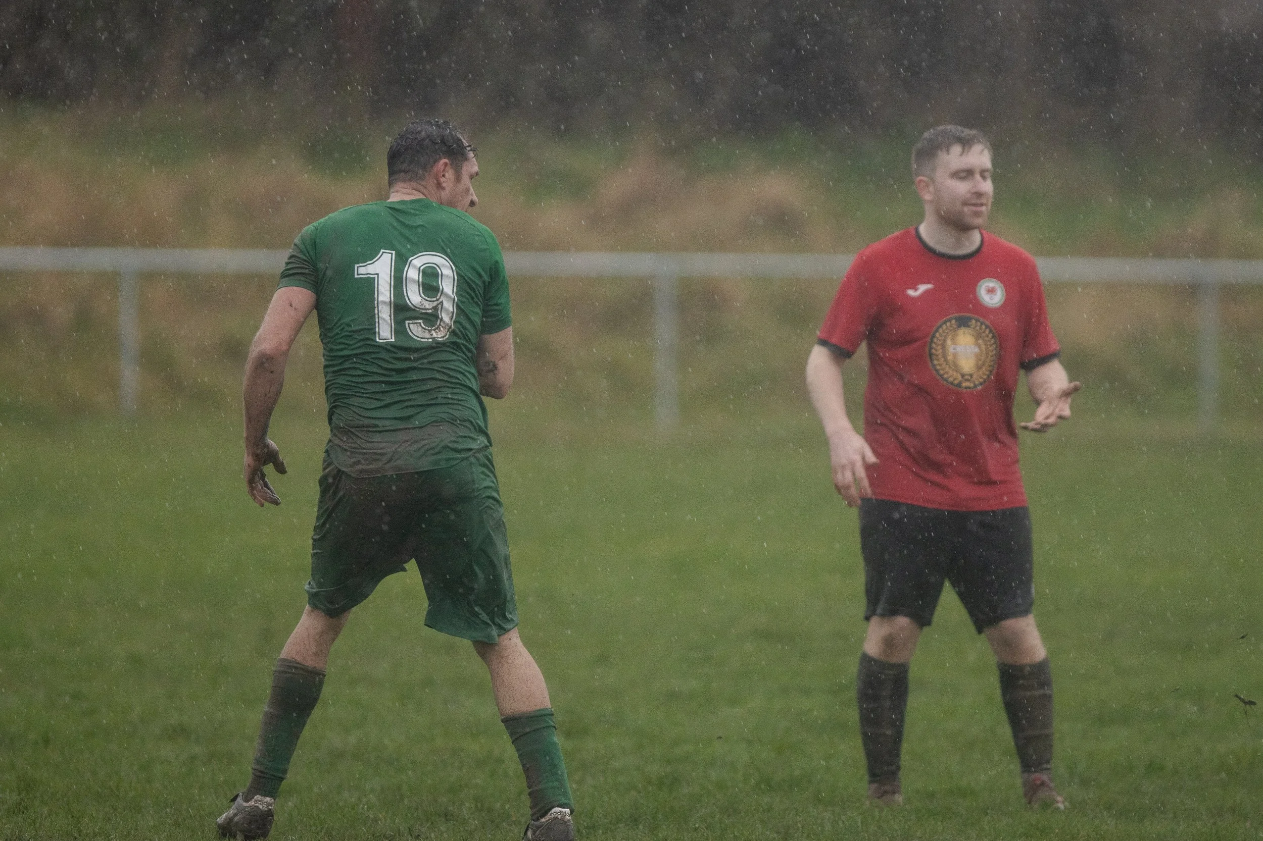 Two male soccer players on a field in the rain. One is wearing a green jersey with the number 19, and the other is in a red jersey with a logo. They appear wet and muddy.