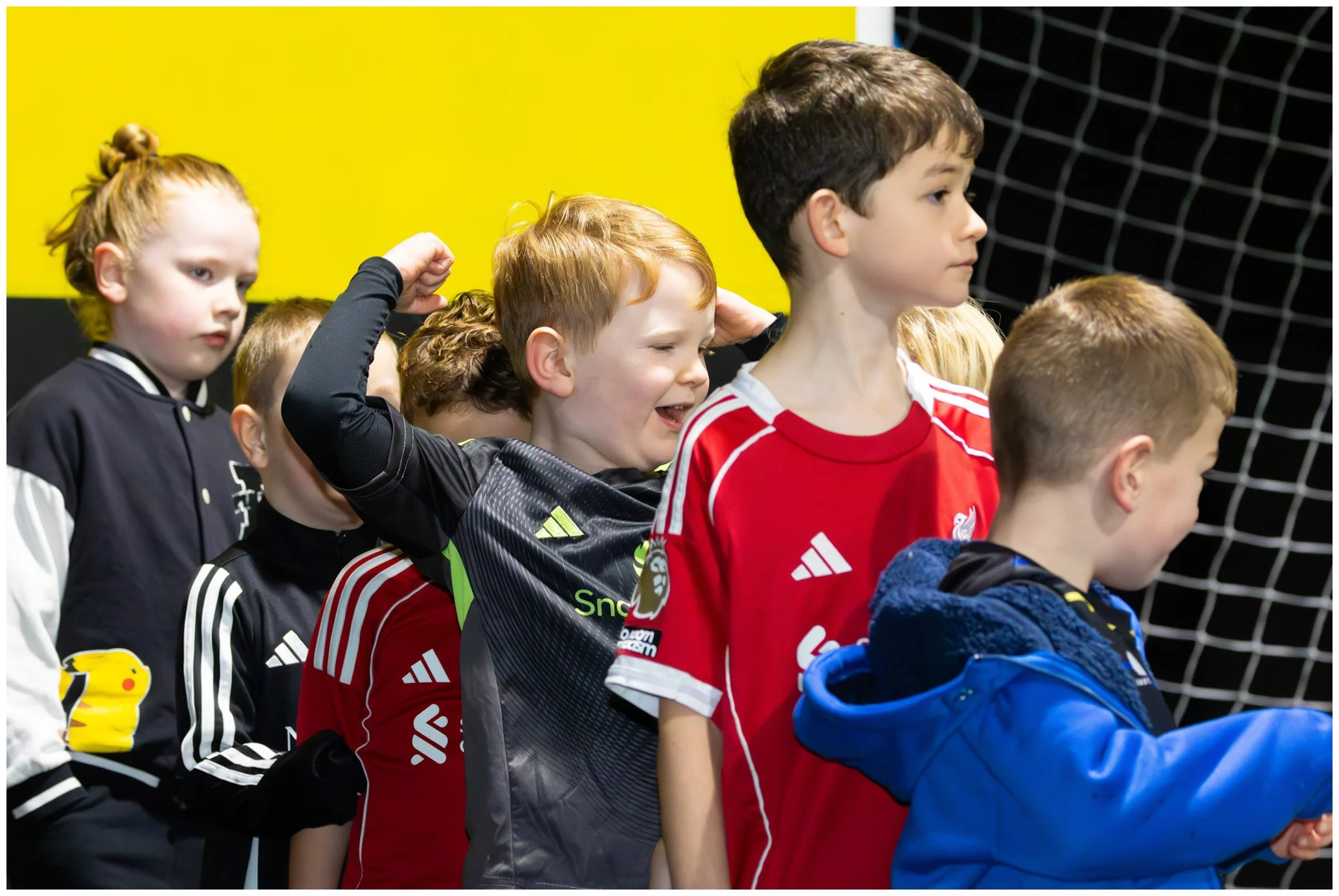 Group of children standing in line inside a sports facility near a goal net, wearing sports jerseys and casual clothes.