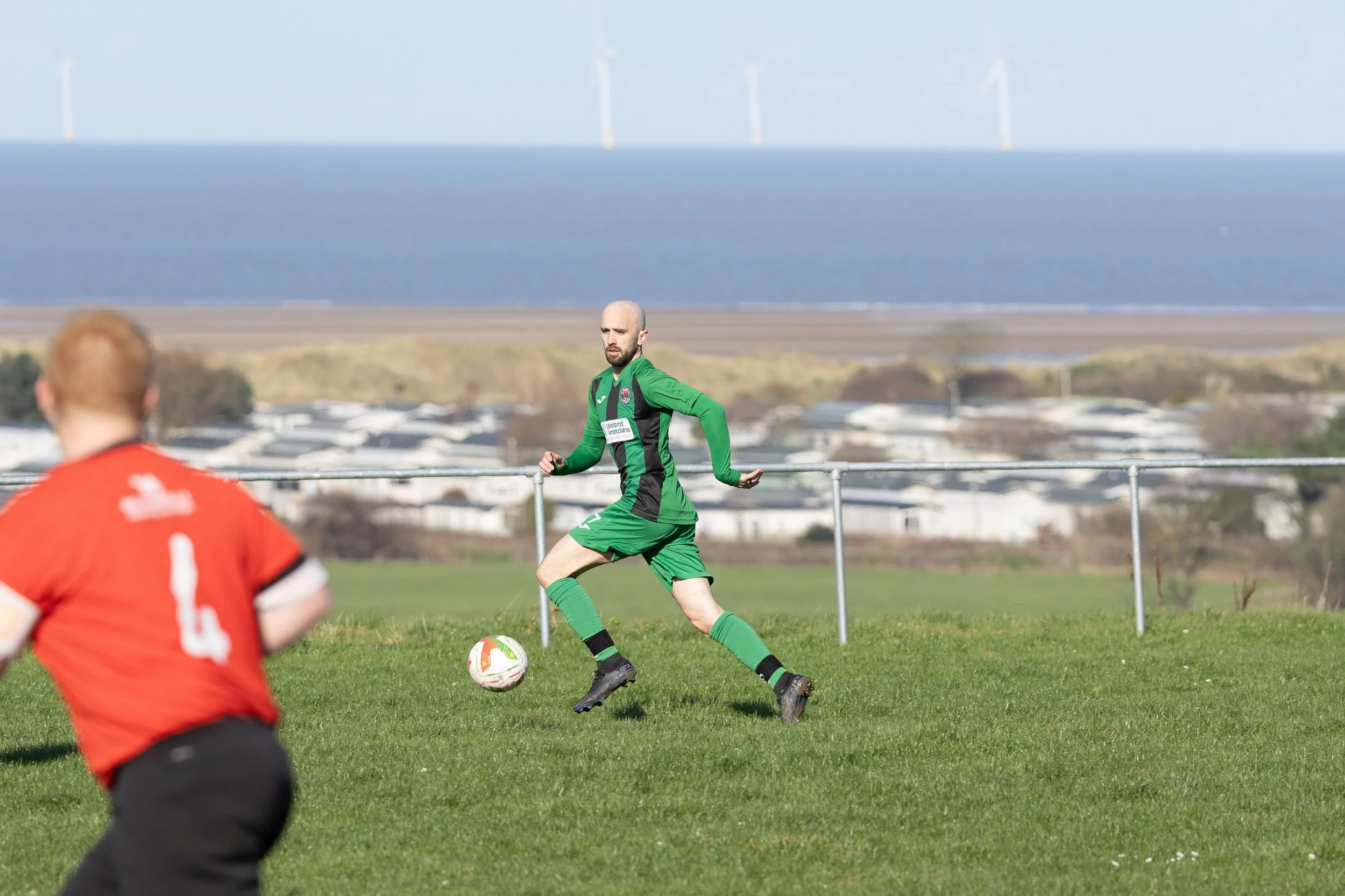 A soccer game on a grassy field with a player in a green uniform about to kick the ball. In the foreground, a player in a red jersey is visible from behind. In the background, there is a view of water and wind turbines under a clear sky.