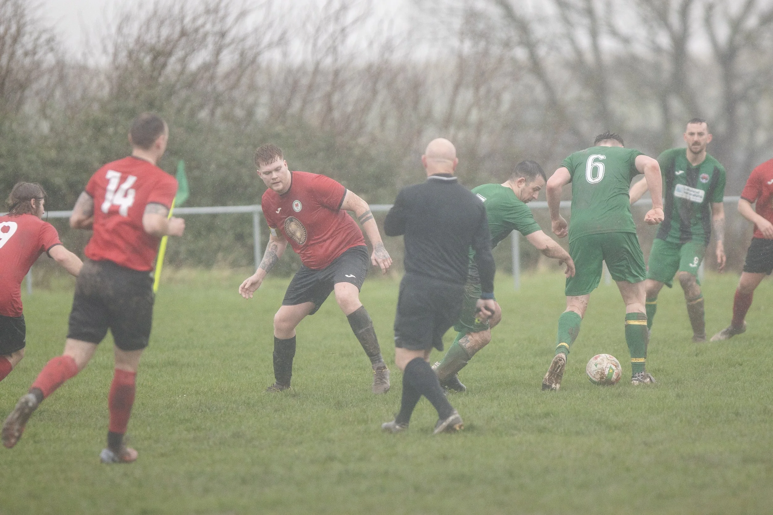 Soccer game with players on a rainy field, some in red and others in green jerseys, with a referee observing.
