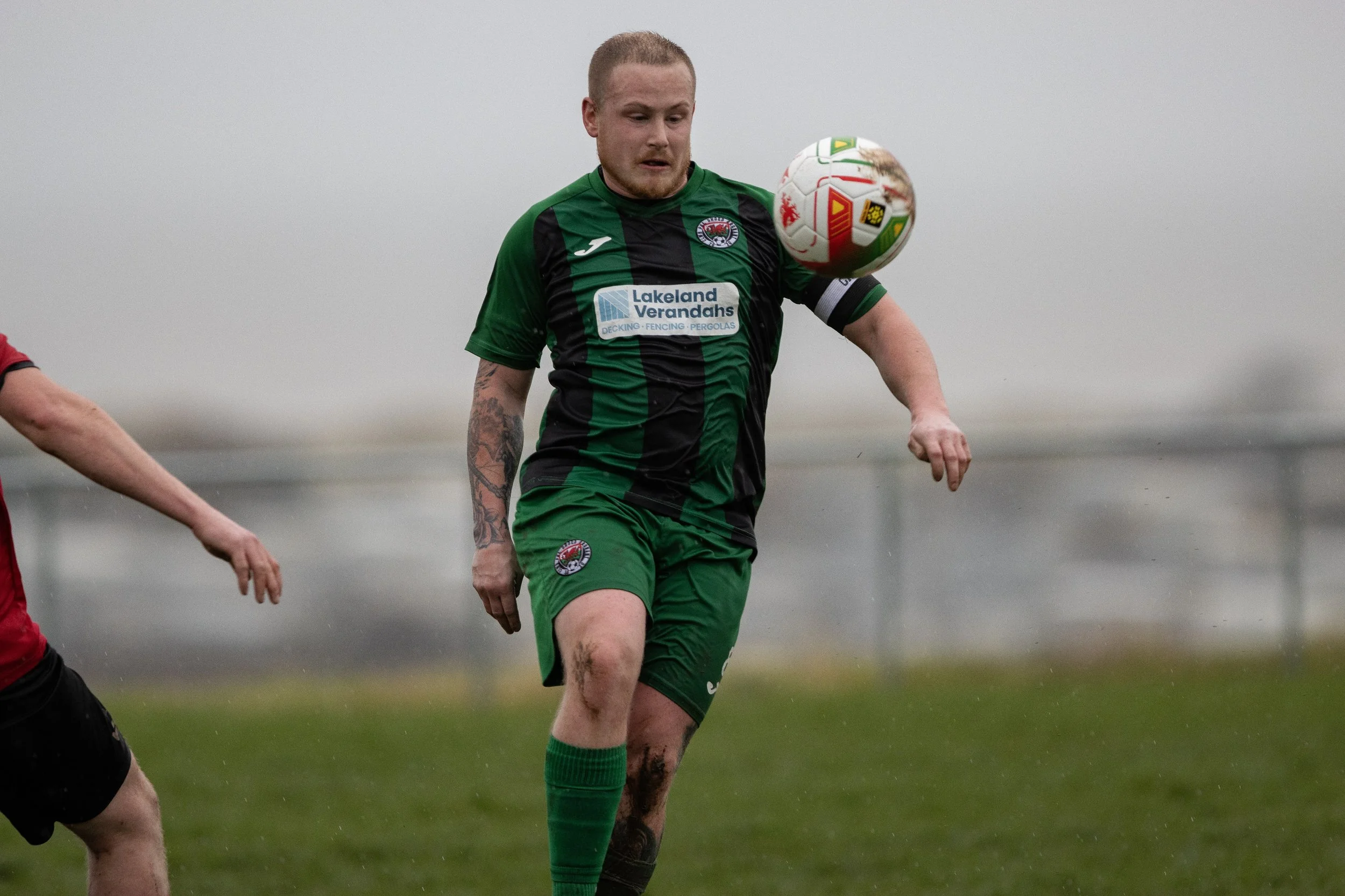 A soccer player in a green and black jersey is heading a soccer ball during a game on a rainy day, with a cloudy sky in the background.