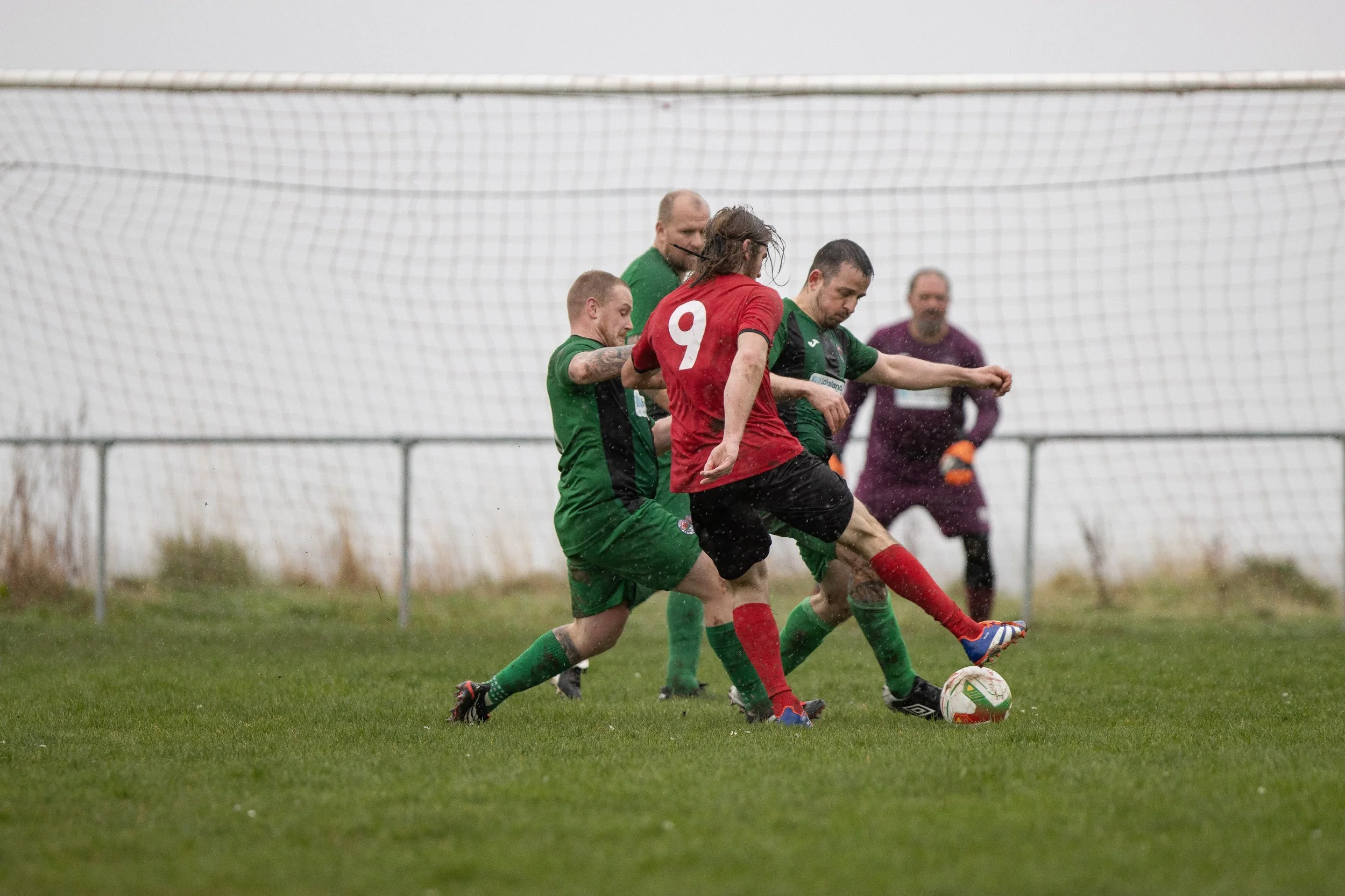 Four men playing soccer on a wet field, with a goal in the background. Two players are wearing green jerseys, one in a red jersey with the number 9, and a goalkeeper in purple.