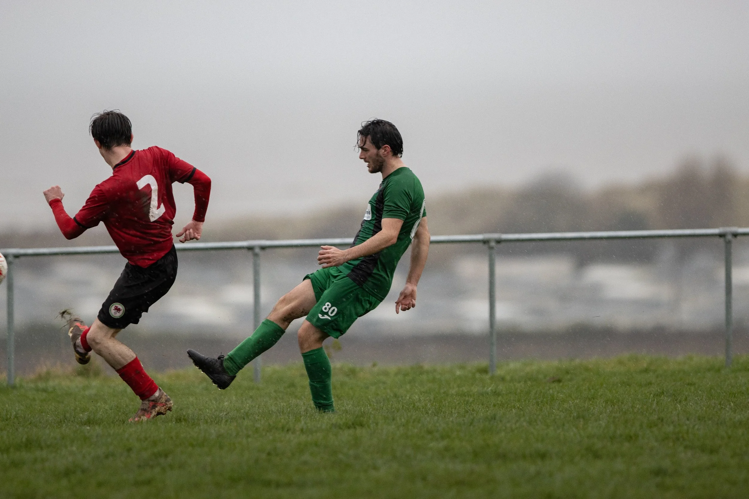 Two soccer players in red and green uniforms competing for the ball on a grassy field, with a cloudy sky in the background.