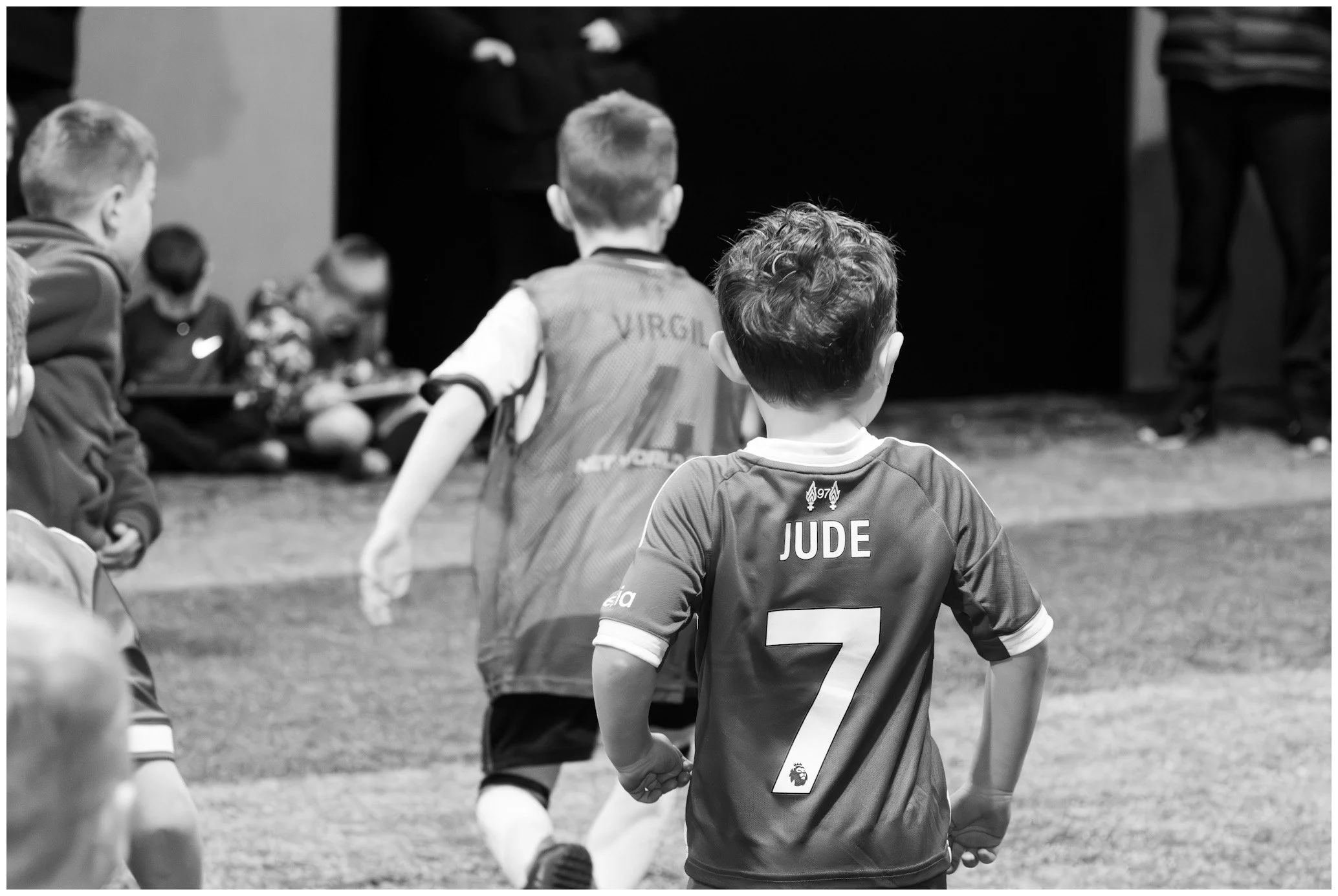Children playing soccer, with one child in the foreground wearing a jersey with the name 'JUDE' and number 7, and another child in the background wearing a jersey with the word 'VIRGIL' visible on the back.