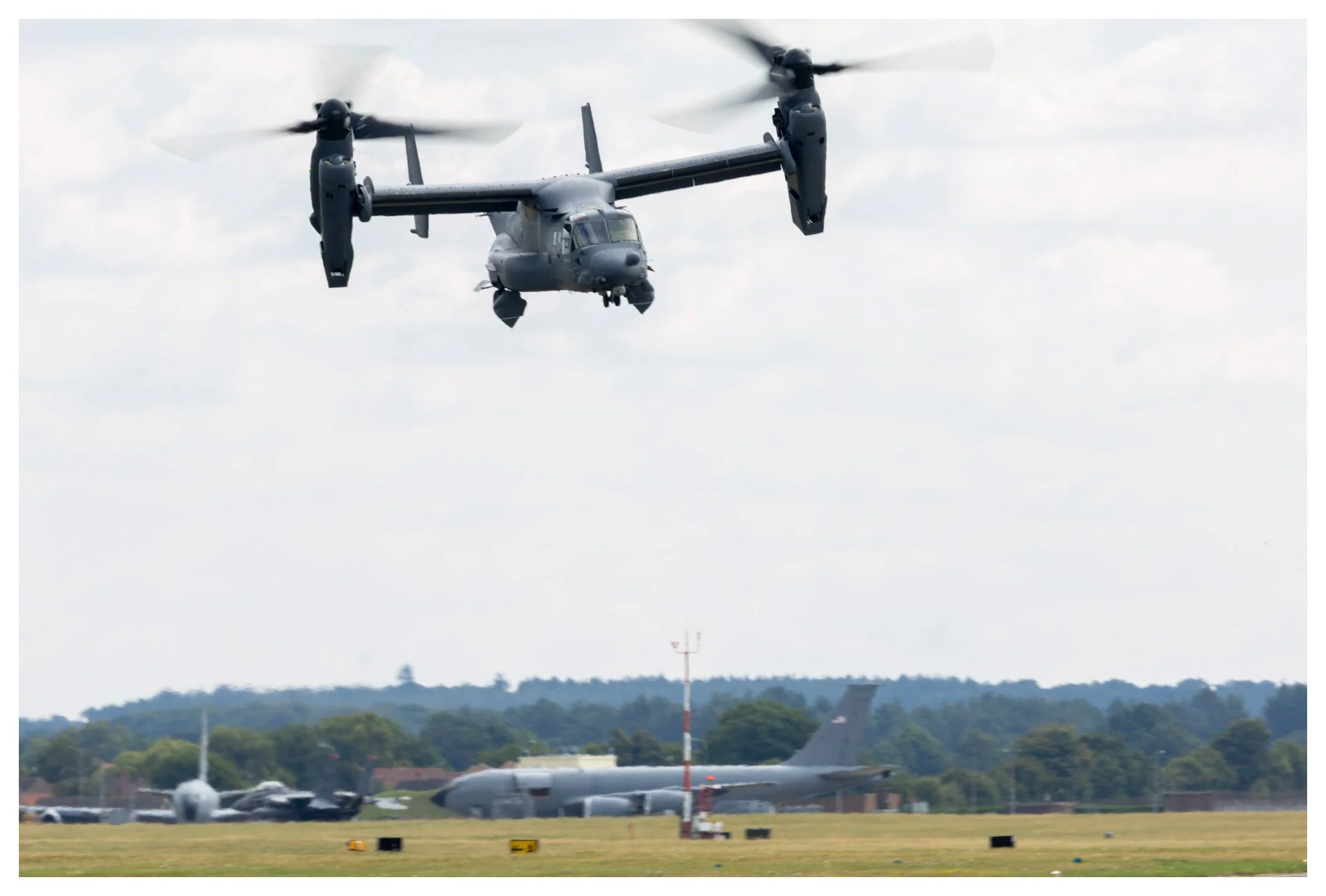 A military tiltrotor aircraft taking off or landing at an airfield with other aircraft and trees in the background.