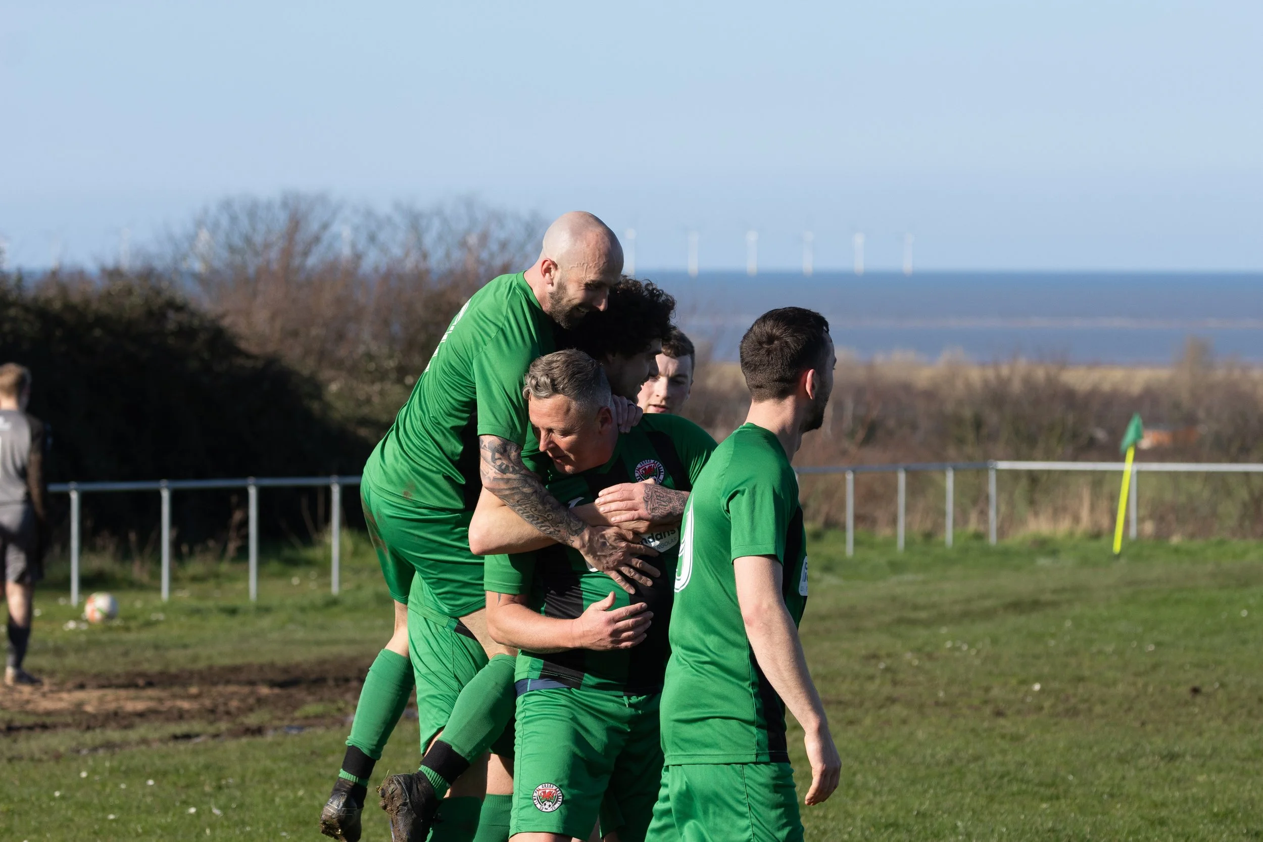 Soccer players in green uniforms celebrating a goal on the field, with a border fence and windswept landscape in the background.
