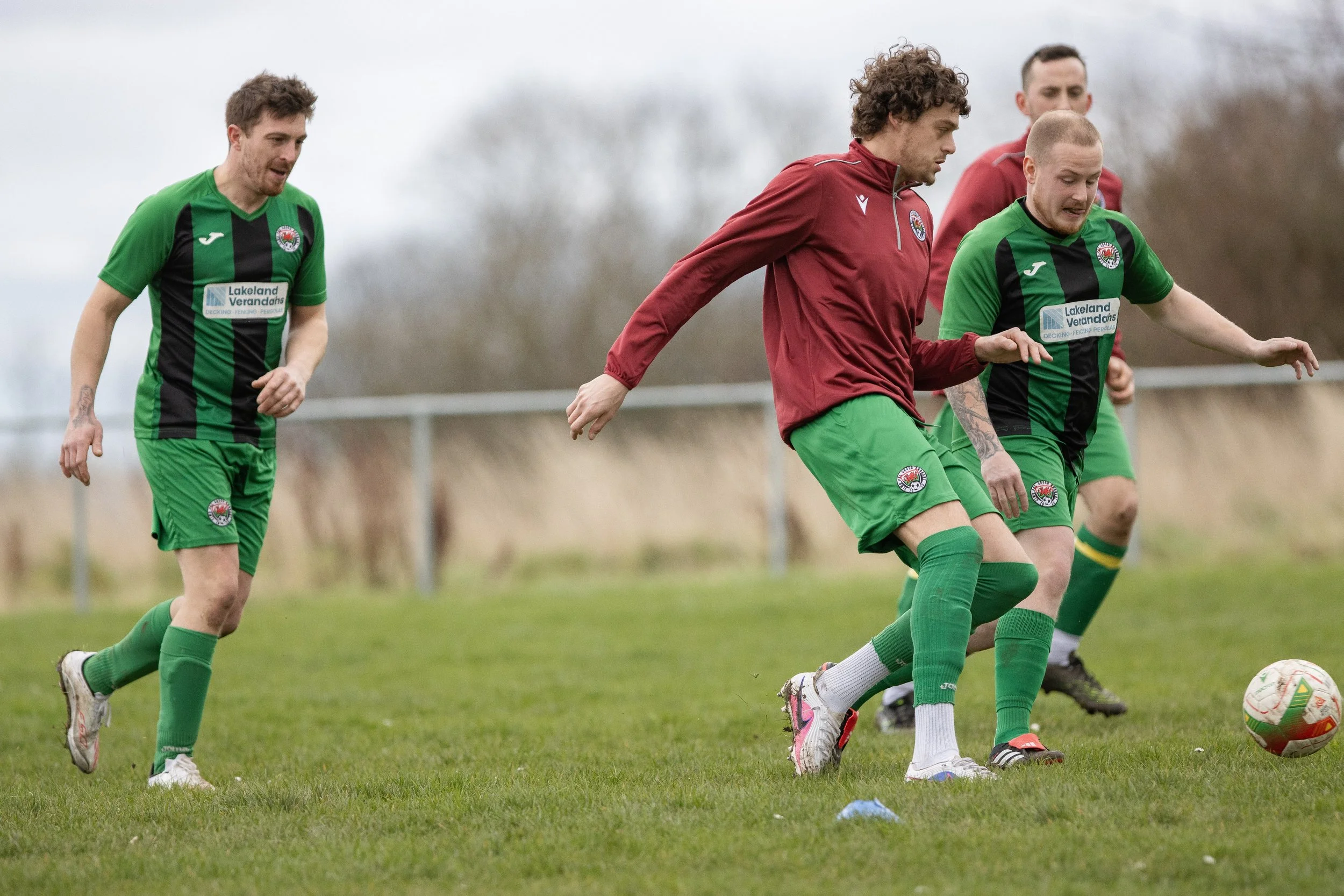 Four soccer players on a grass field, two wearing green and black uniforms and two wearing red and green, competing for the ball during a match.