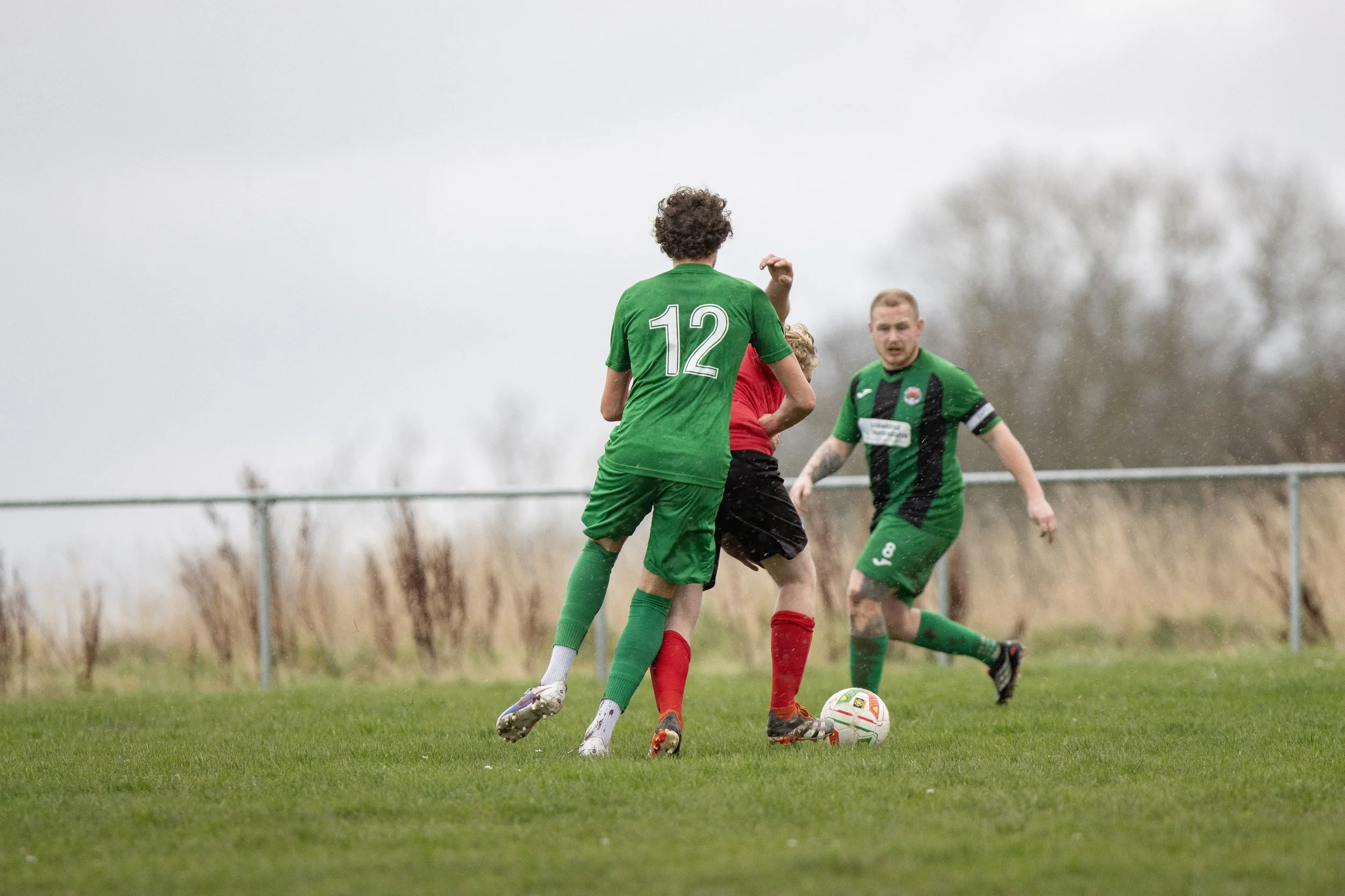 Three soccer players in green and red jerseys compete for the ball on a grassy field.