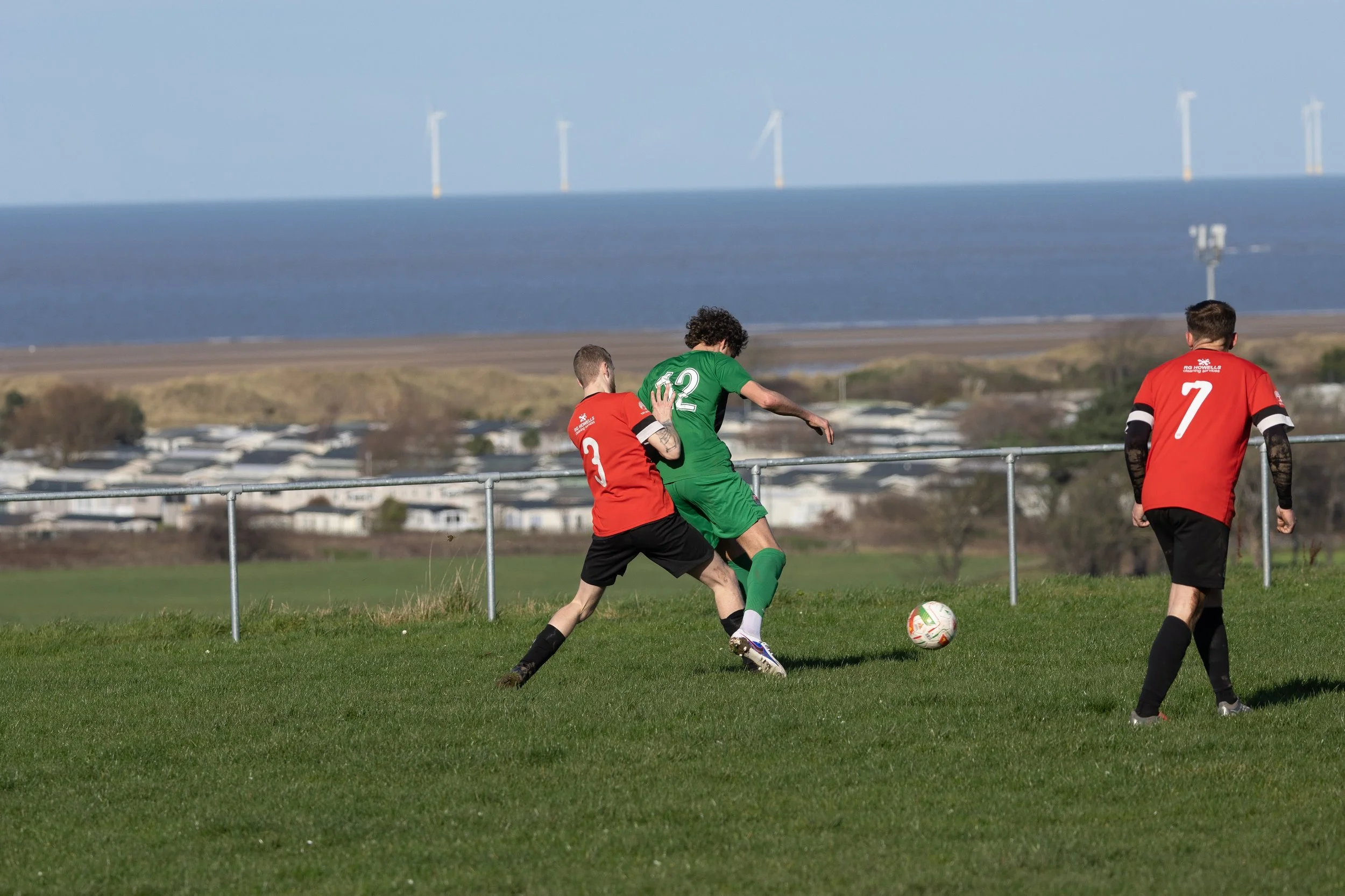 Three soccer players on a green field, two in red jerseys and one in a green jersey, competing for the ball with a coastal landscape and wind turbines in the background.