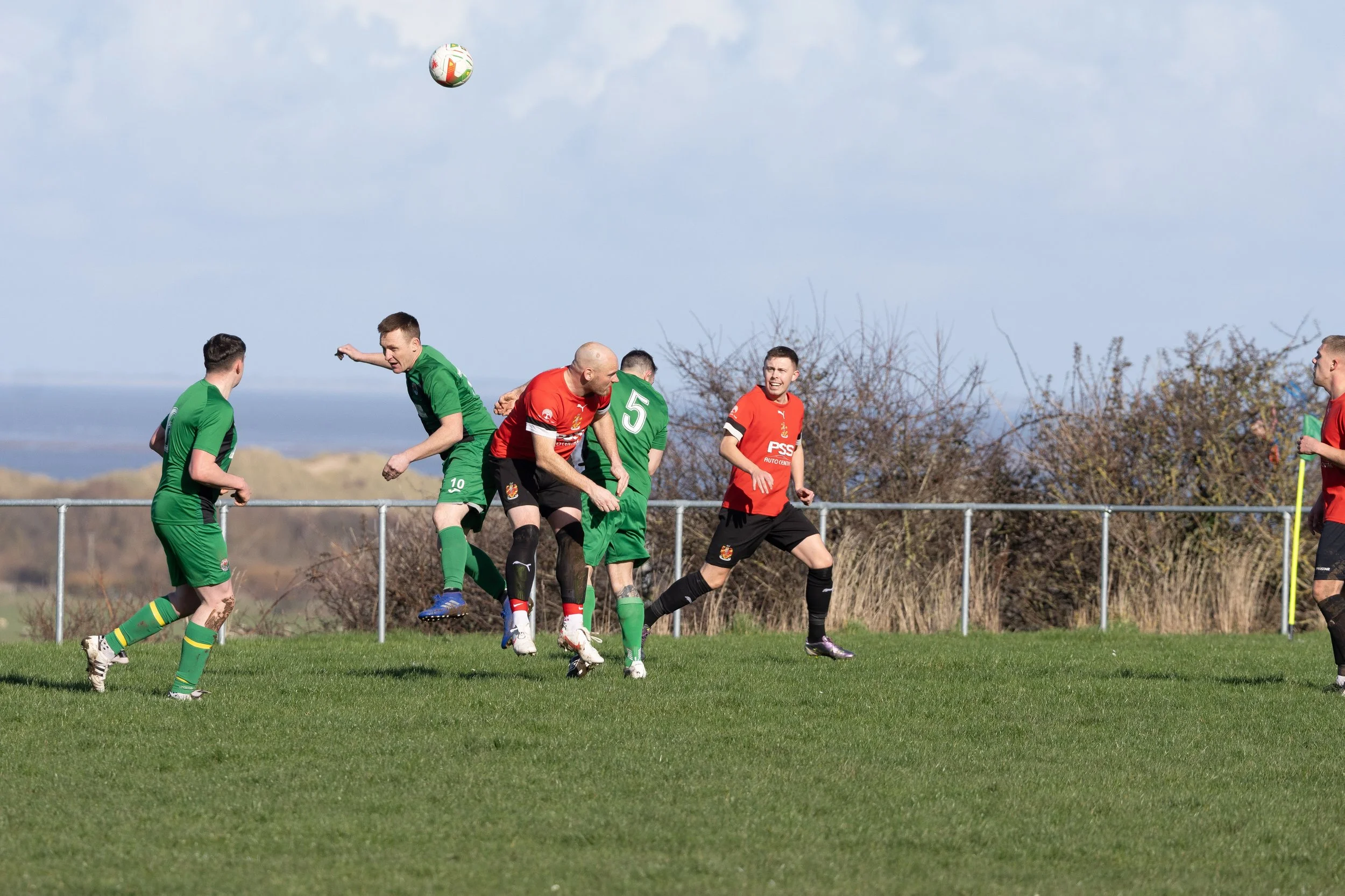 Soccer players in green and red jerseys competing for the ball during a match on a grassy field with a fence and trees in the background.