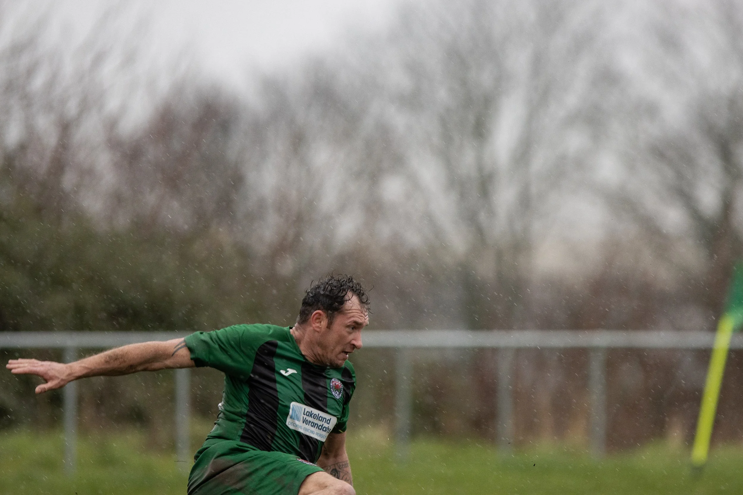 A man in a green and black sports jersey with a sponsor logo, running or falling on a rainy outdoor field, with blurred trees and a yellow corner flag in the background.