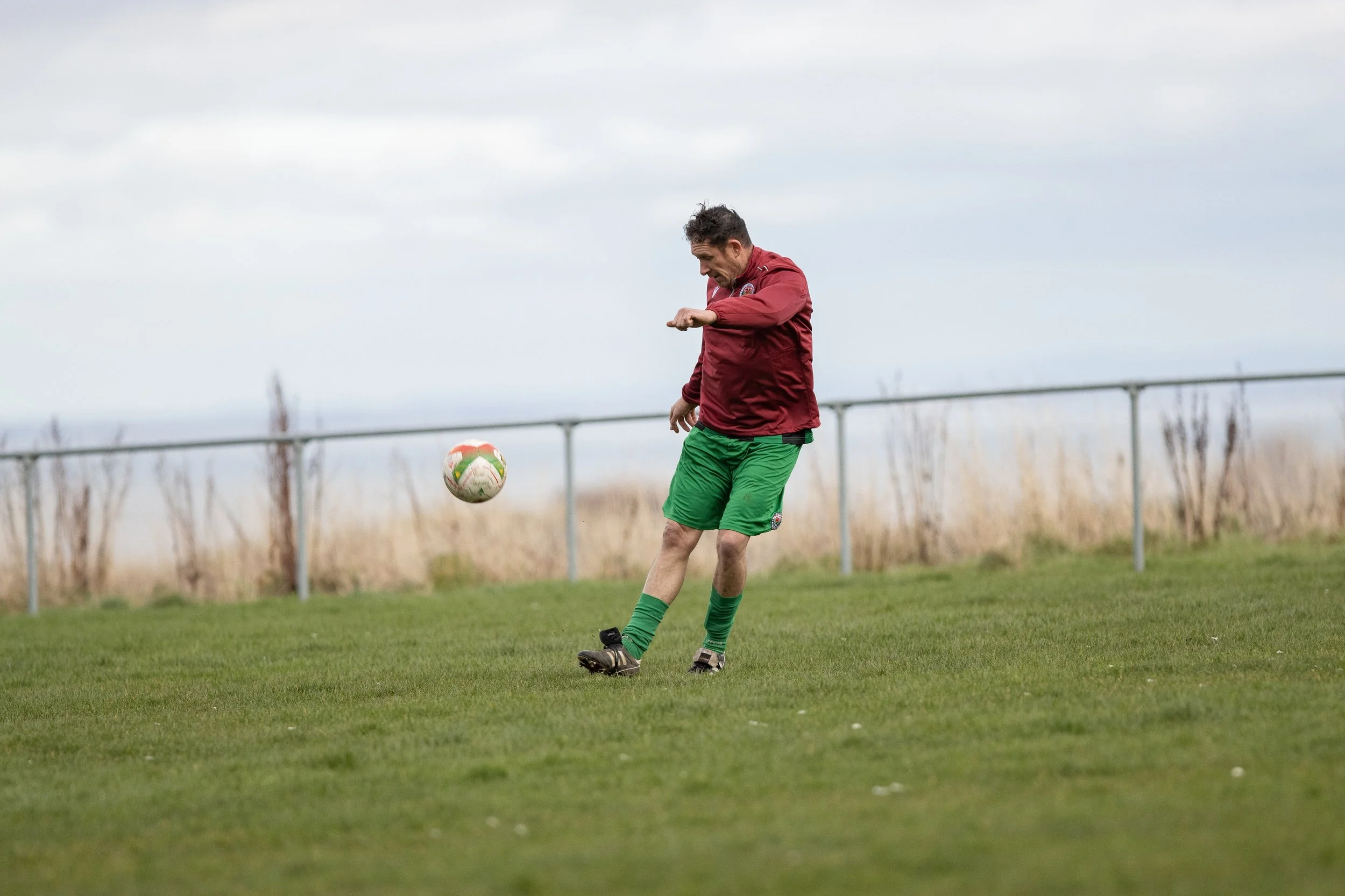 A man in a red jacket and green shorts kicks a soccer ball on a grassy field, with a bare fence and cloudy sky in the background.