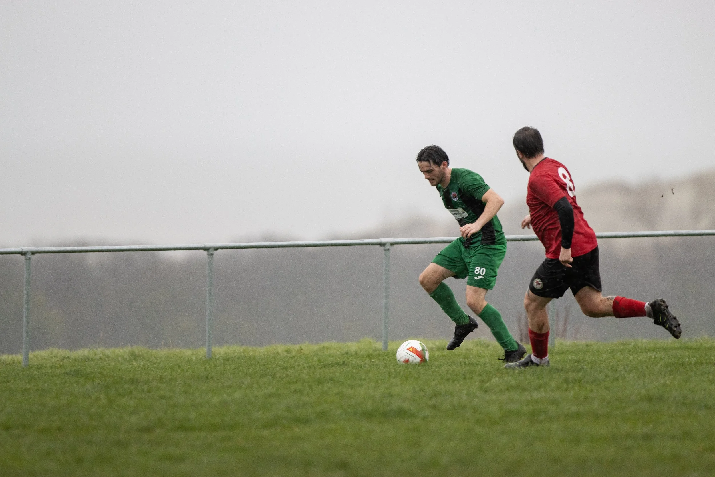 Two soccer players, one in green and one in red, competing for the ball on a grassy field on a rainy day.