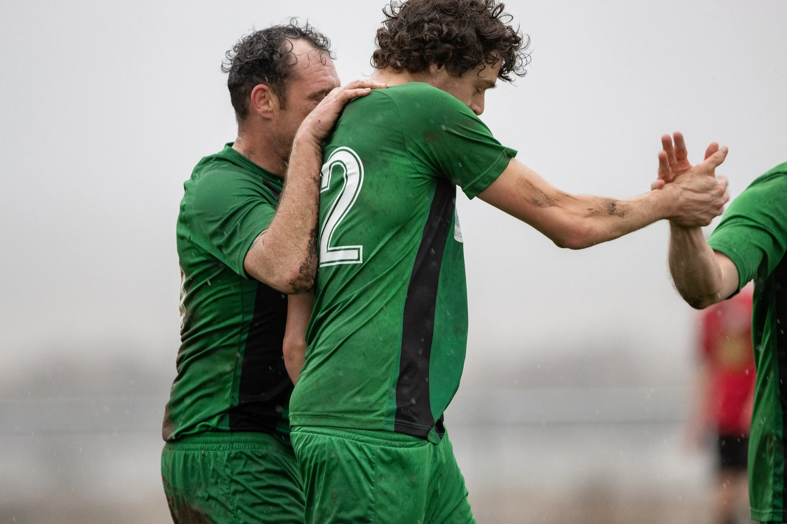 Two soccer players in green uniforms exchanging a high five on a rainy day, one embracing the other from behind.