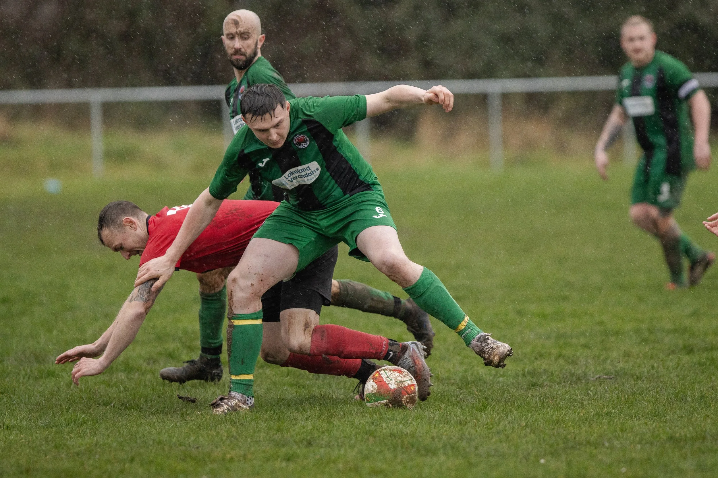 Soccer players in green and red uniforms competing for the ball on a wet grass field during a rainy match.