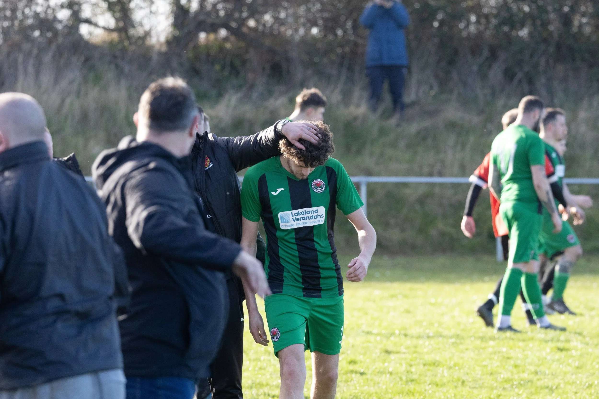 A soccer player in green uniform being comforted by a coach on the field after a game, with other players and spectators in the background.