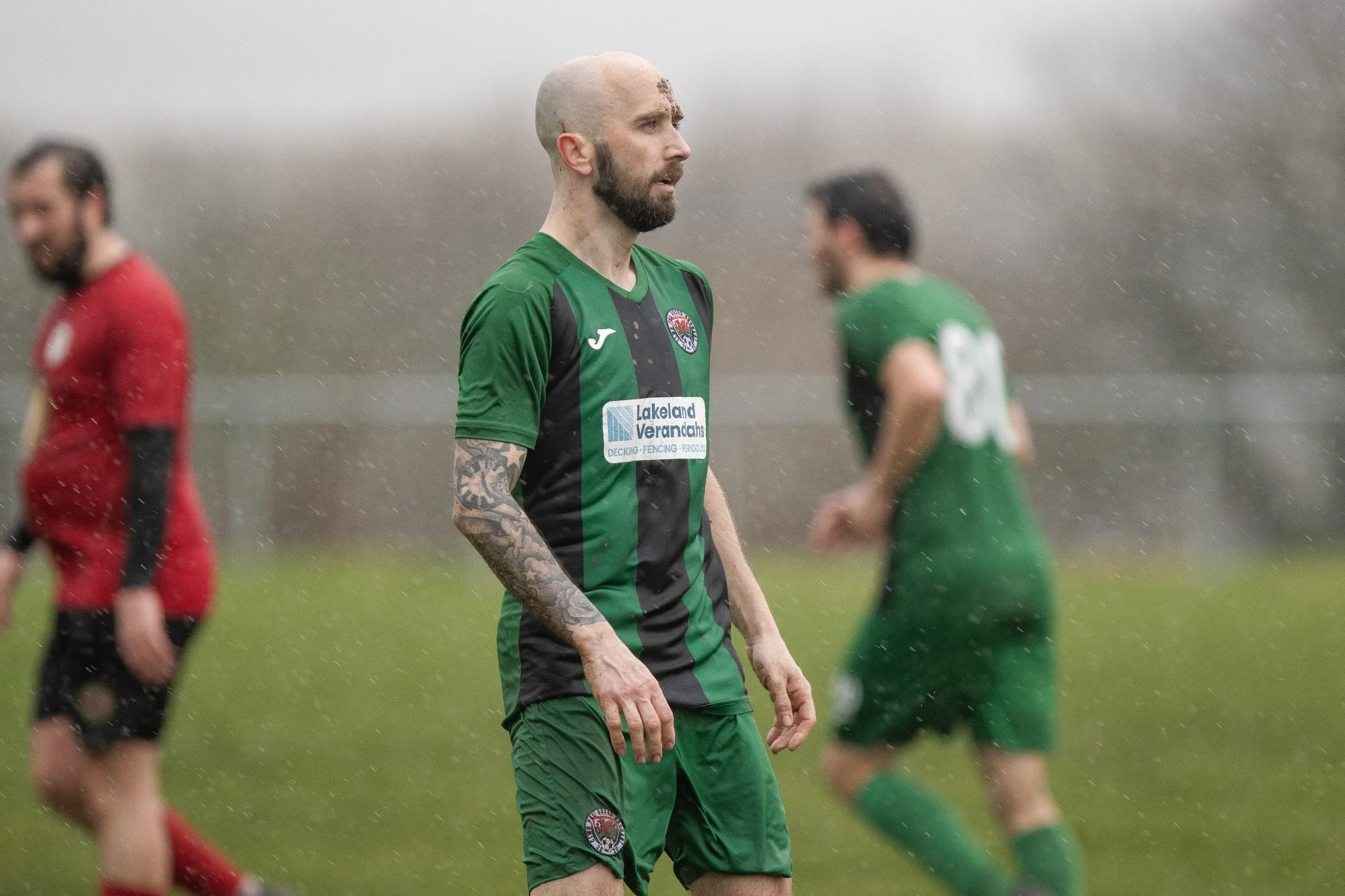 Soccer players on a field in the rain, with one player in a green and black uniform in the foreground, and others in red and green uniforms in the background.